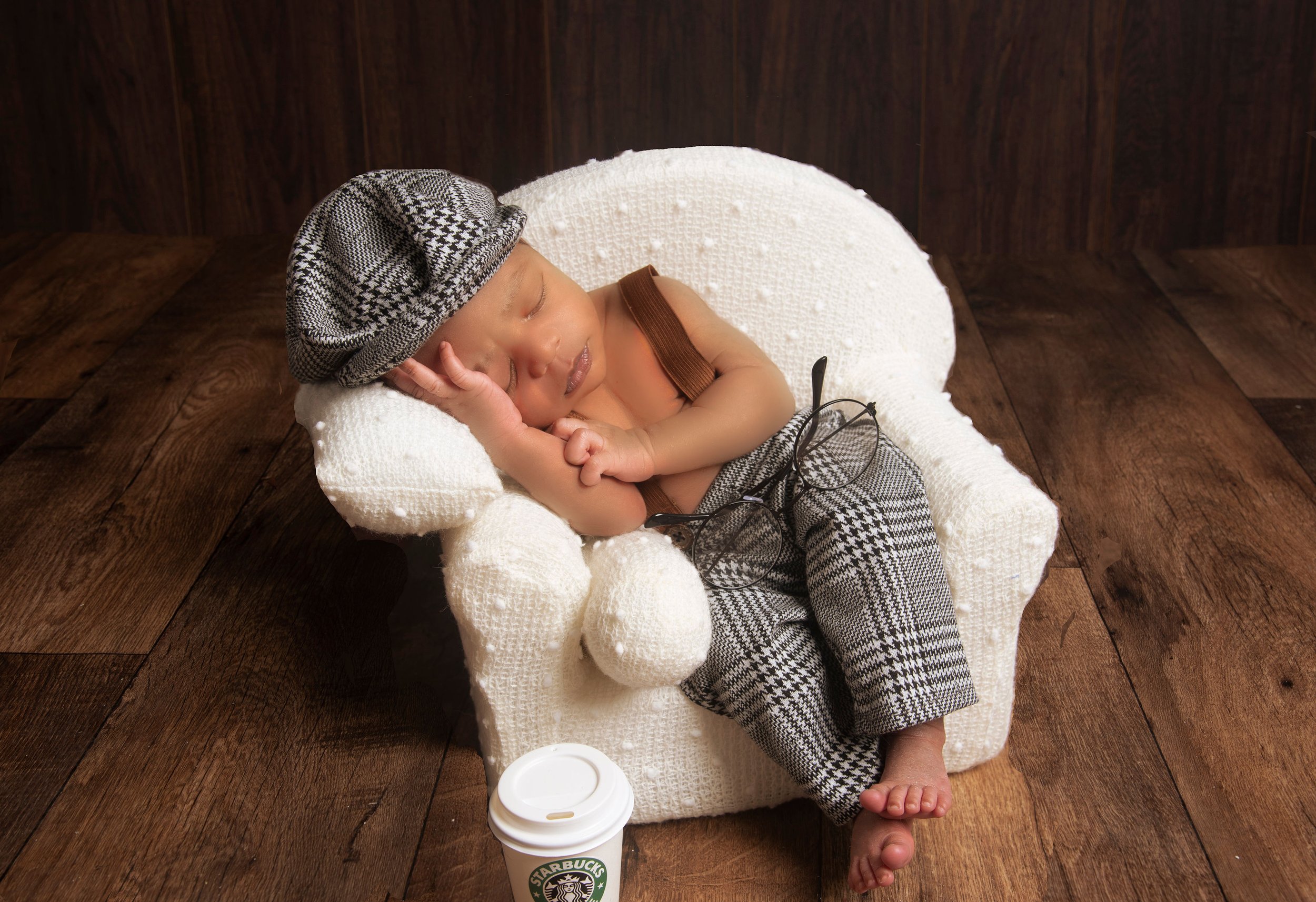 A sleeping baby wearing a patterned cap and pants, resting on a small white armchair, with glasses on the lap and a Starbucks cup on the floor, on a wooden surface.