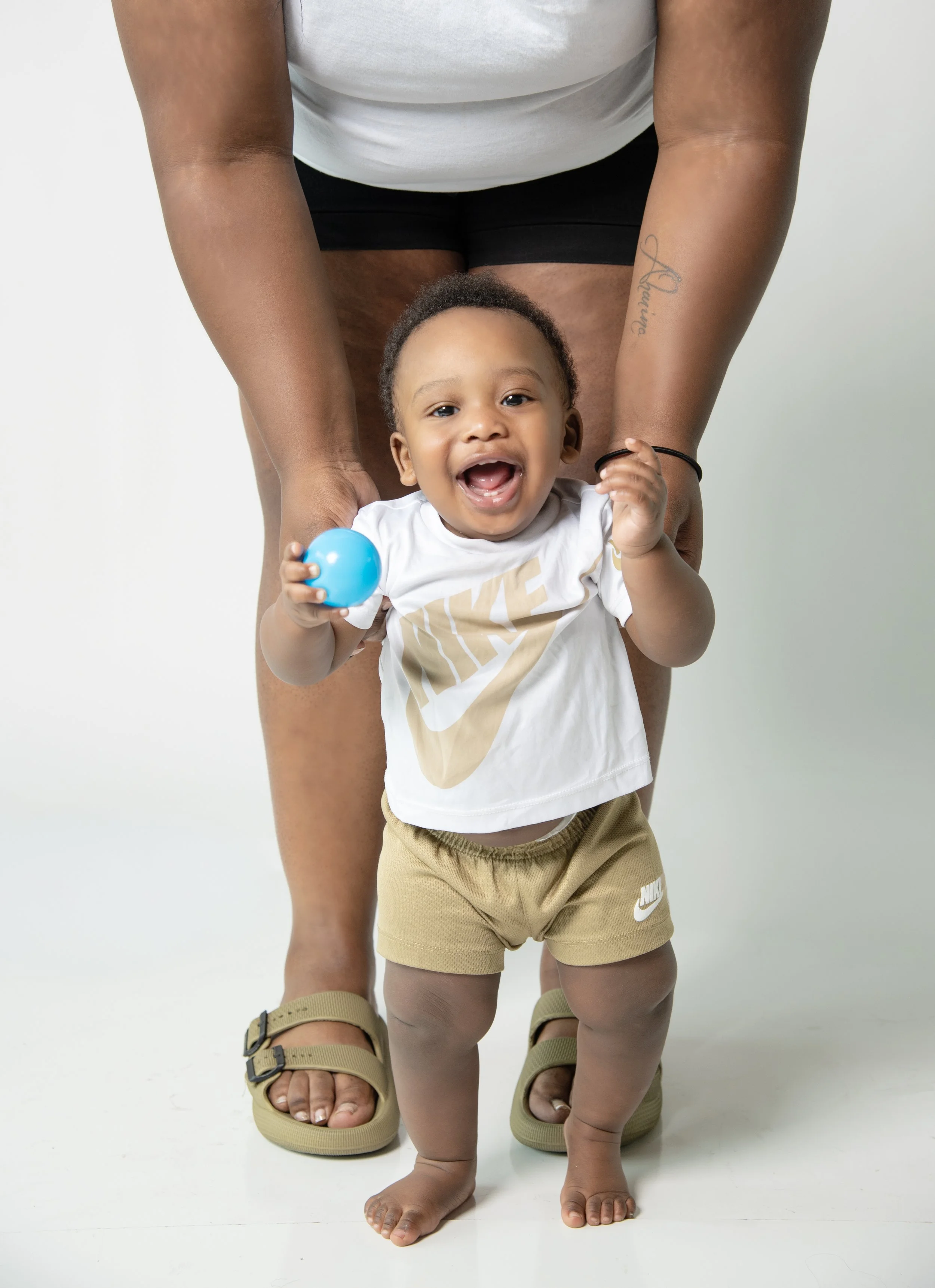 A smiling toddler walking towards the camera, holding a small blue ball in one hand, with an adult bending down behind, holding the child's hands for support. The child is wearing beige Nike shorts and a white Nike t-shirt, and the adult is wearing a
