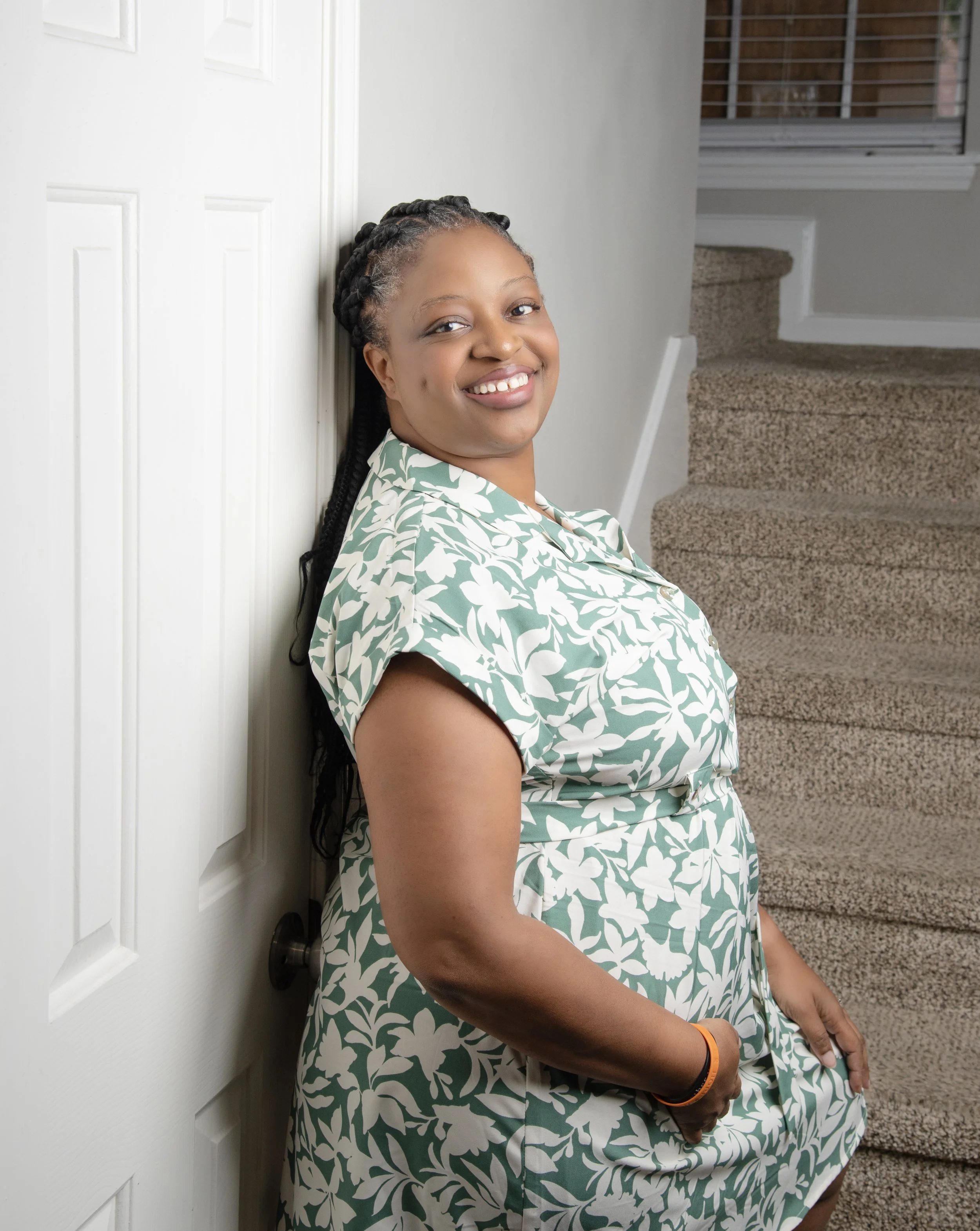 Patricia Johnson, owner of PDJ Photography smiling leaning against a white doorframe on a staircase, wearing a green and white patterned dress.