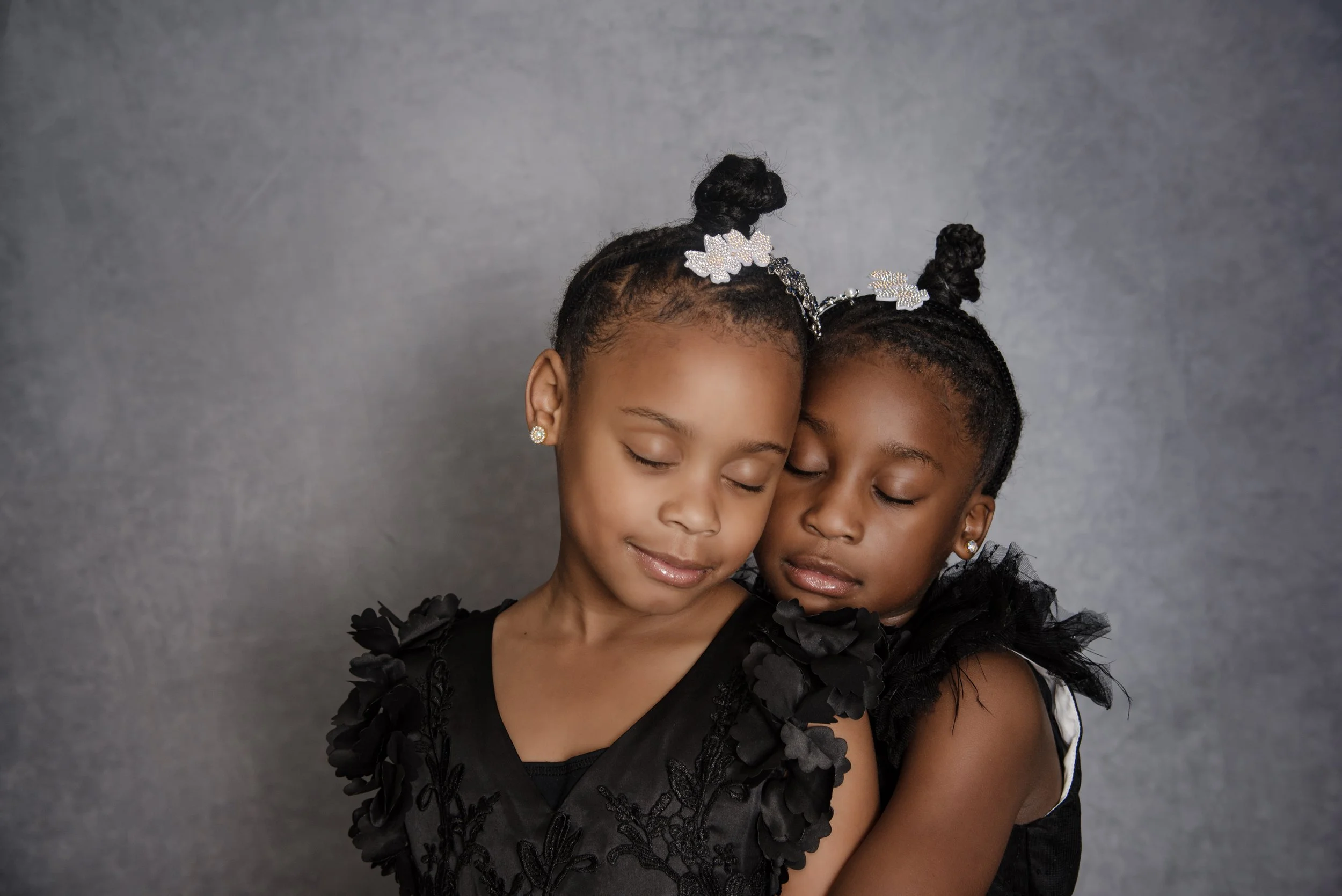 Two young girls with closed eyes and serene expressions, hugging each other, dressed in black dresses with floral details and wearing matching hair accessories against a plain gray background.