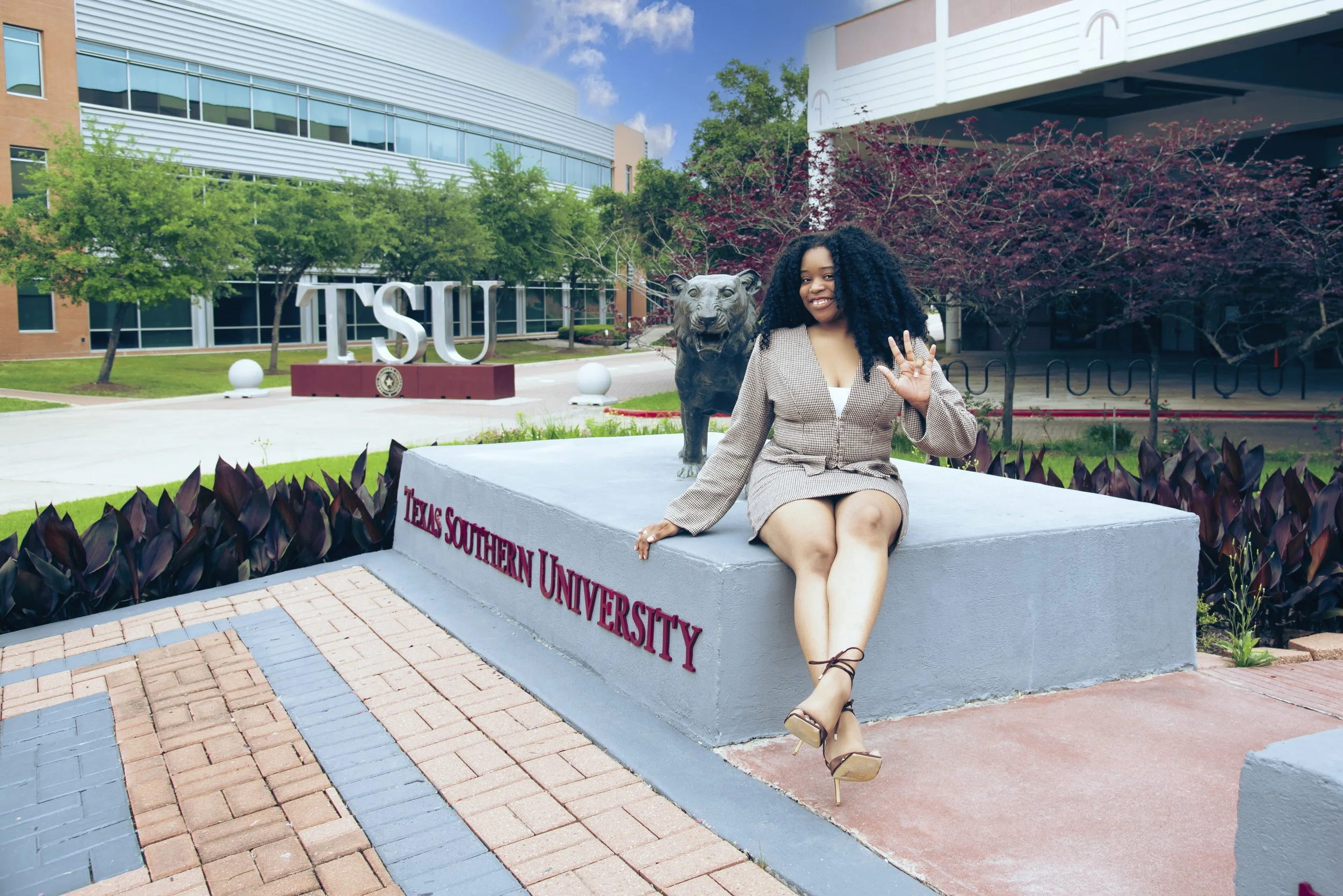 A woman sitting on a concrete platform with a lion statue behind her, in front of a university building with a sign reading 'TSU' and 'TEXAS SOUTHERN UNIVERSITY', surrounded by trees and bushes.