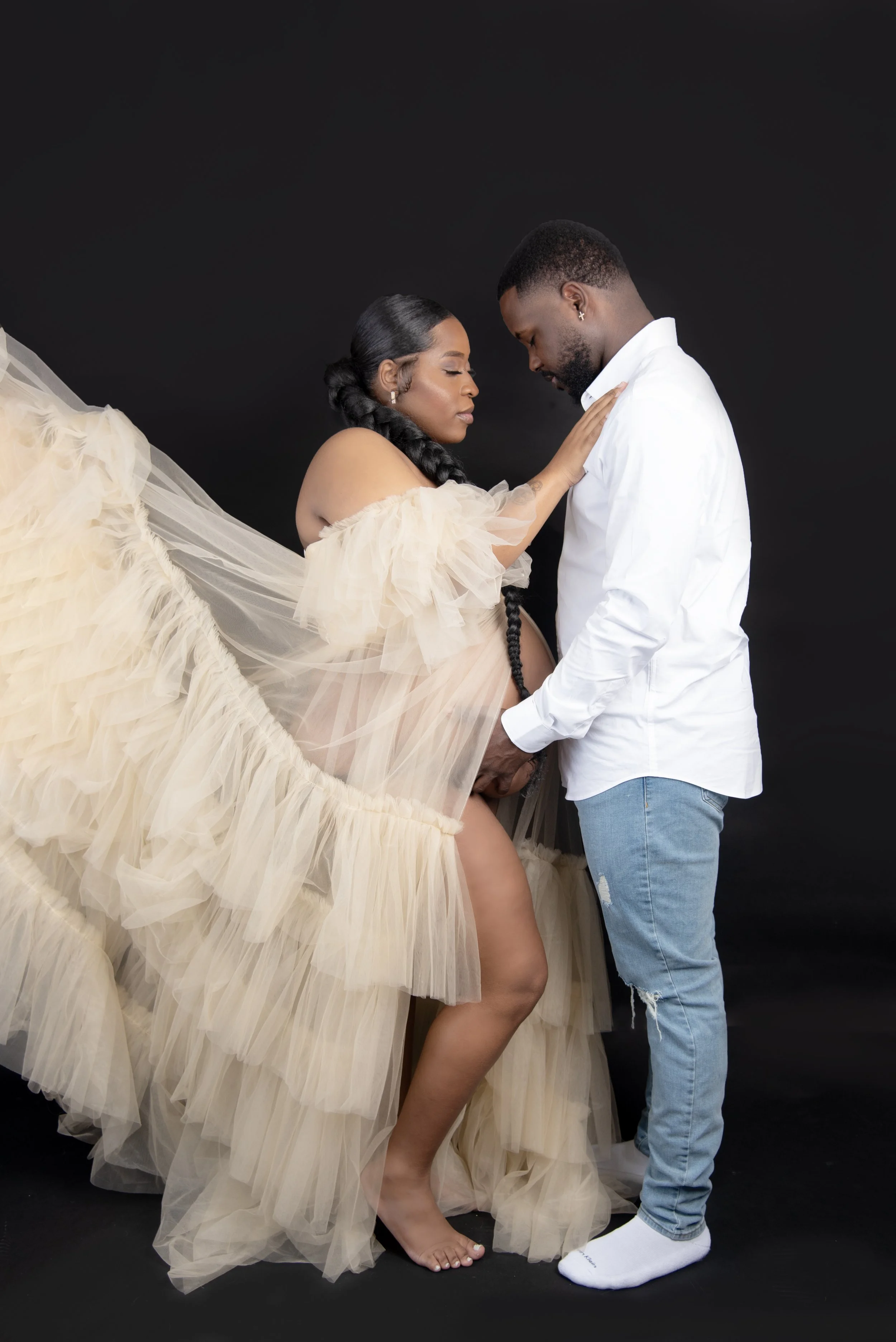 A pregnant woman in a beige, ruffled tulle gown is gently caressing her belly with a man standing close to her, touching her shoulder, against a dark background.