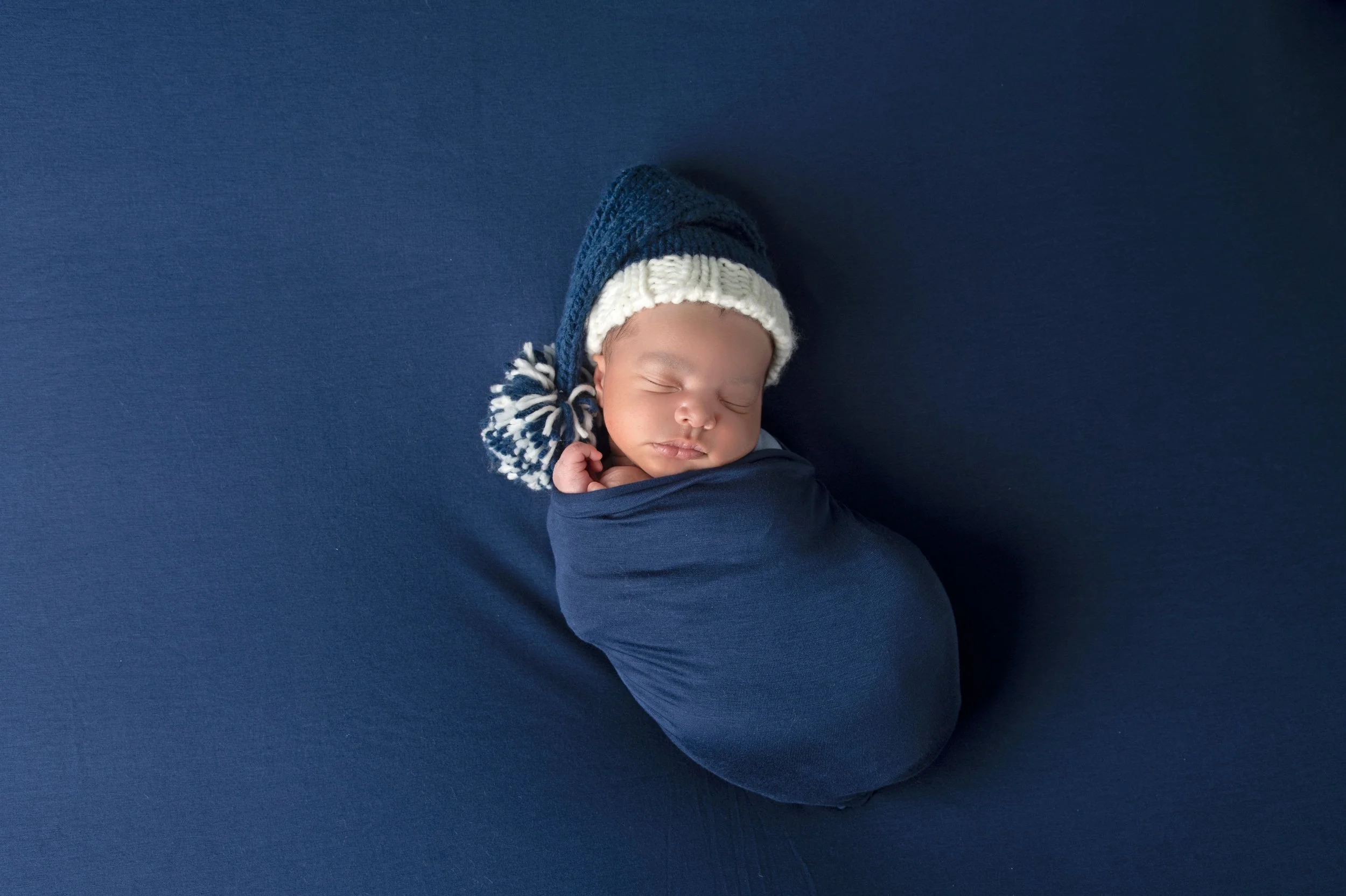 A sleeping baby wrapped in a blue swaddle, wearing a blue and white knit hat with a pom-pom, lying on a solid blue background.
