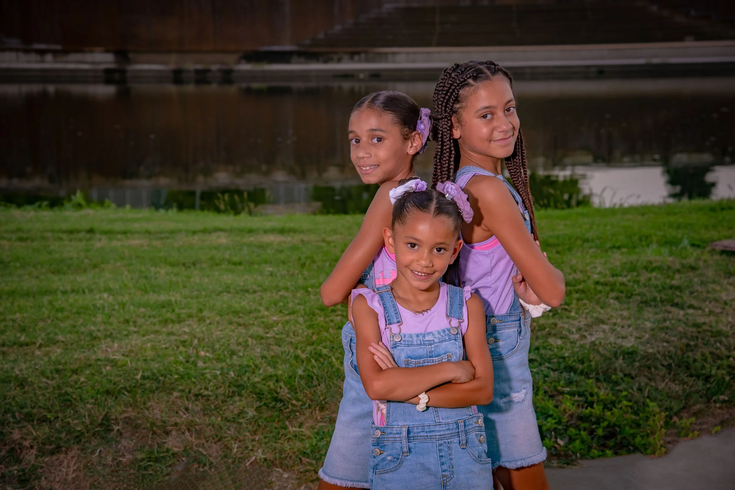 Three young girls with hair in braids and bows, standing on grass near water, smiling at camera.