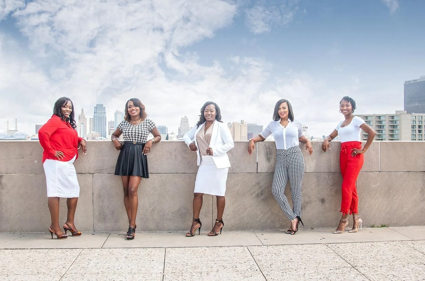 Five realtors standing on a rooftop ledge with a city skyline in the background, dressed in professional and fashionable clothing.
