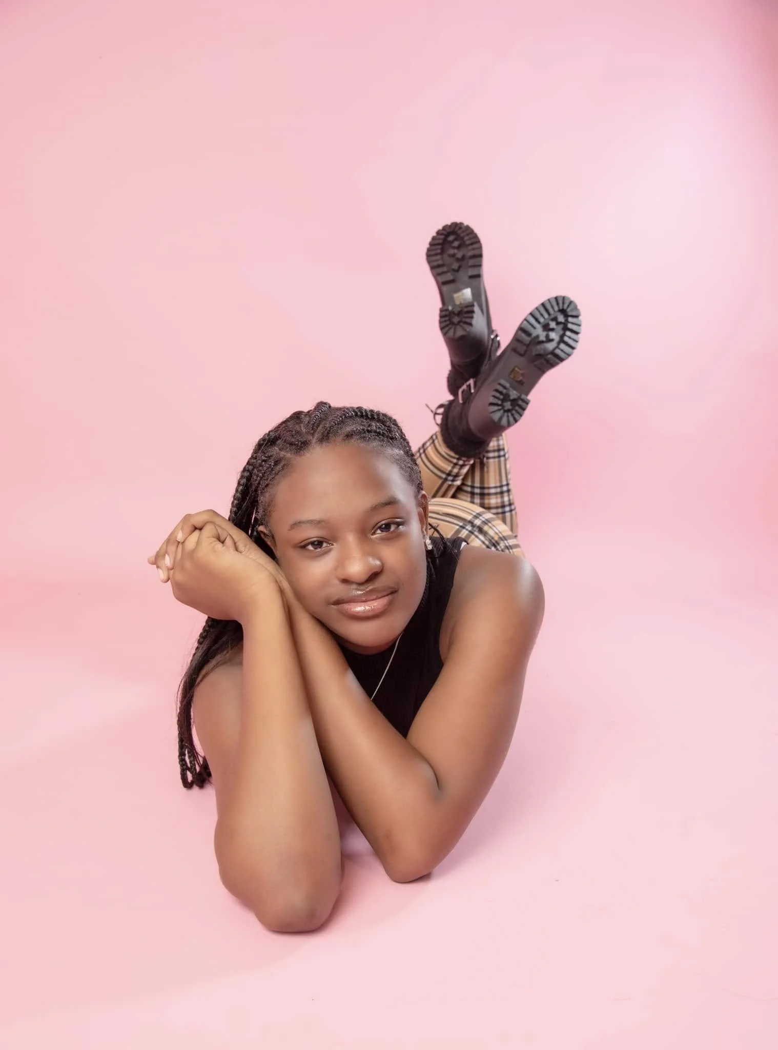 A young woman with braided hair lies on a pink background, resting her head on her hands, smiling slightly, wearing a black sleeveless top, plaid pants, and black boots, with her feet up and crossed.