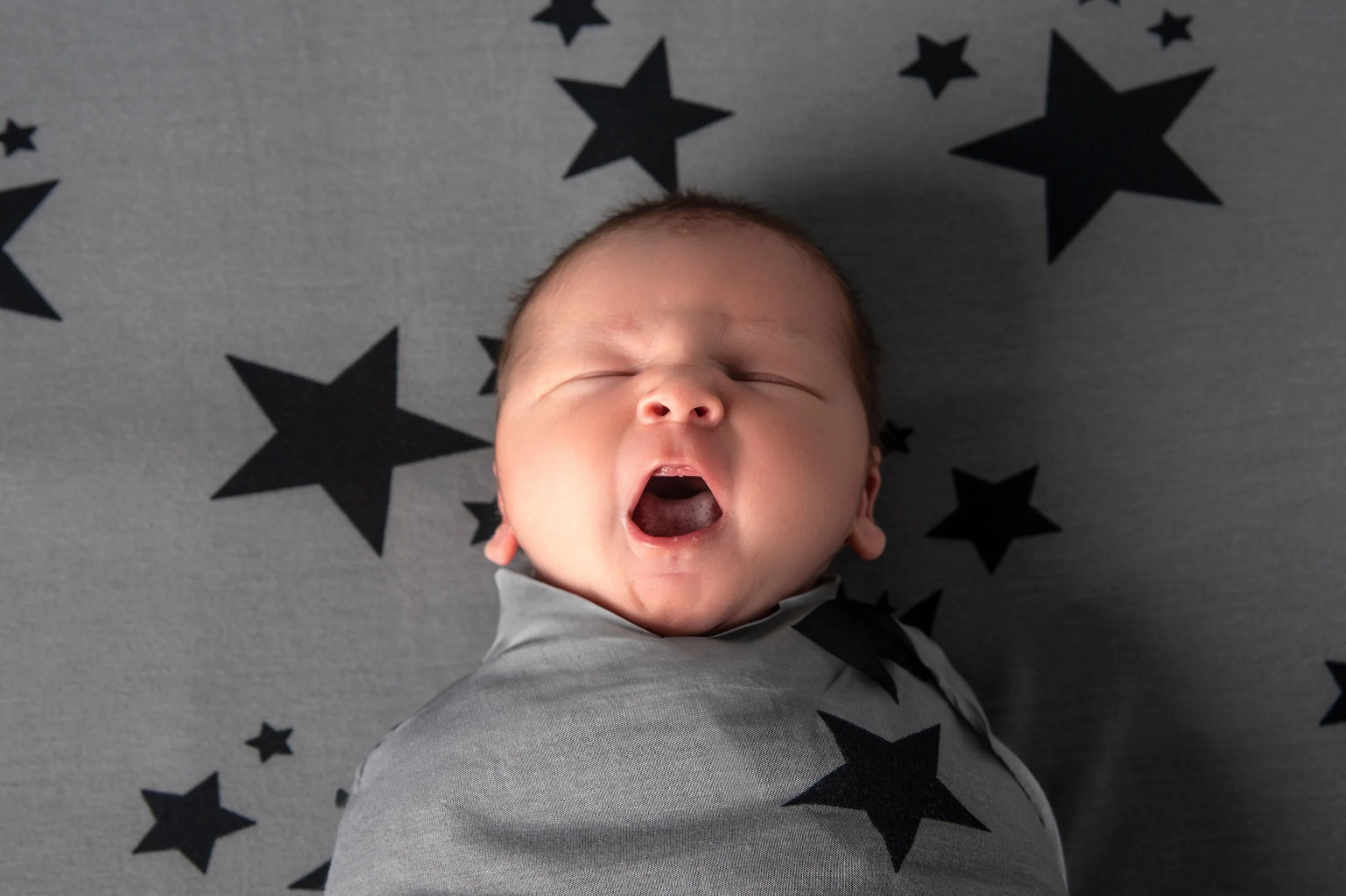 A yawning sleeping baby lying on a gray blanket with black star patterns.