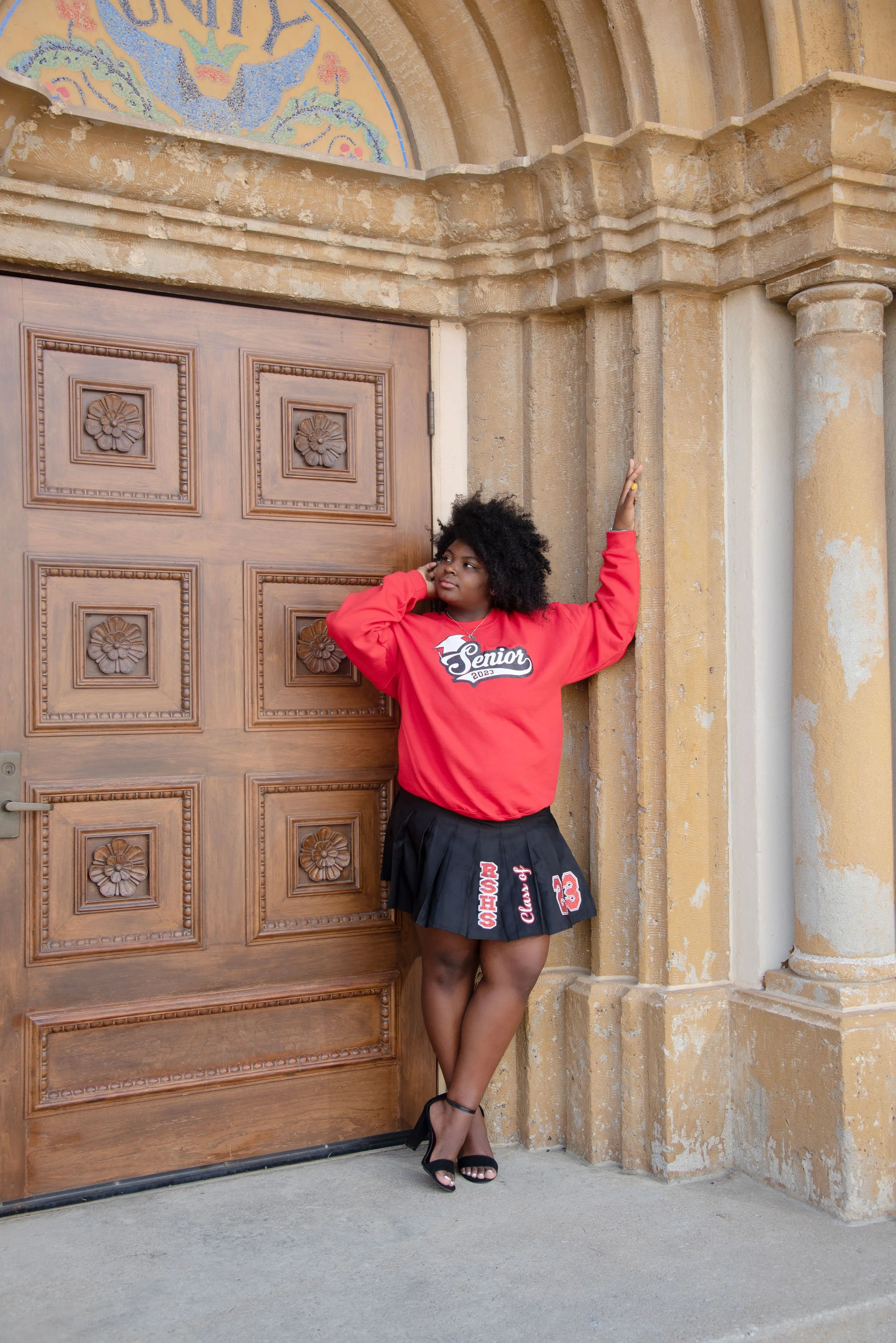 Young woman in a red sweatshirt with 'Senior 2023' on it and a black skirt with a sports theme, standing against an ornate wooden door and stone building with columns.