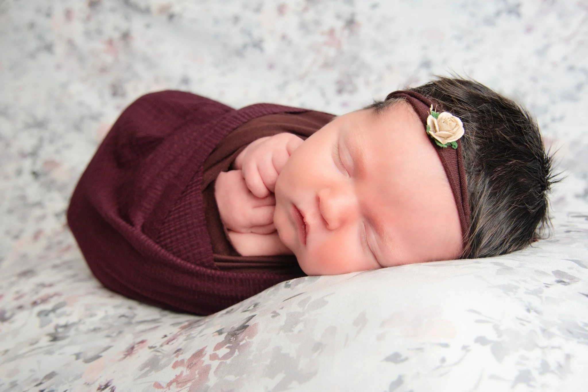 A sleeping newborn baby wrapped in a maroon blanket with eyes closed, laying on a white surface. The baby wears a brown headband with a small cream flower decoration.