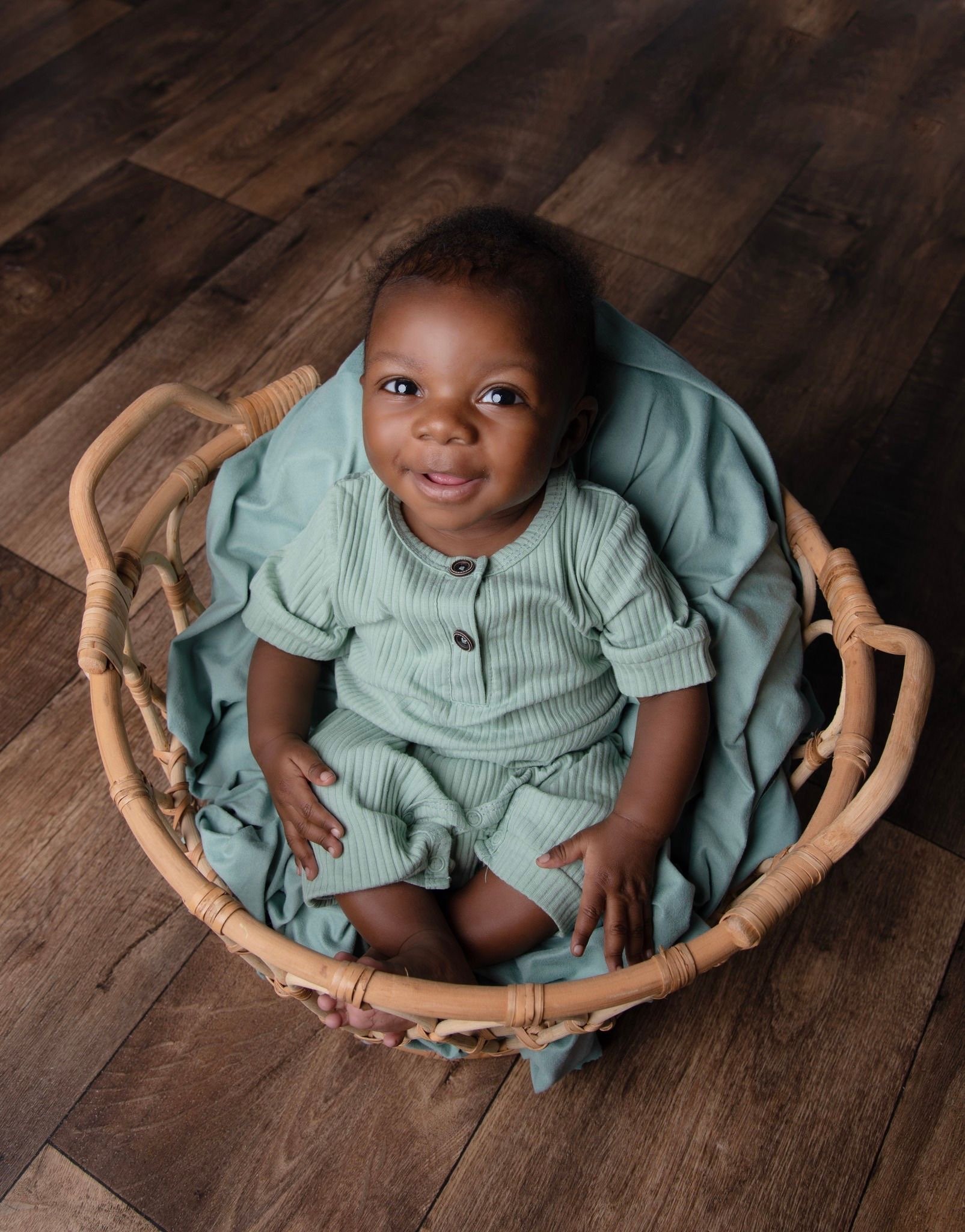 A smiling baby sitting in a wicker basket on a wooden floor, wearing a matching green outfit.