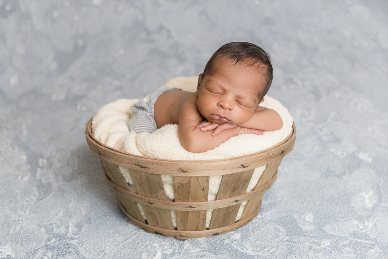 A sleeping newborn baby with dark hair resting on a soft, cream-colored blanket inside a wooden basket on a textured gray surface.