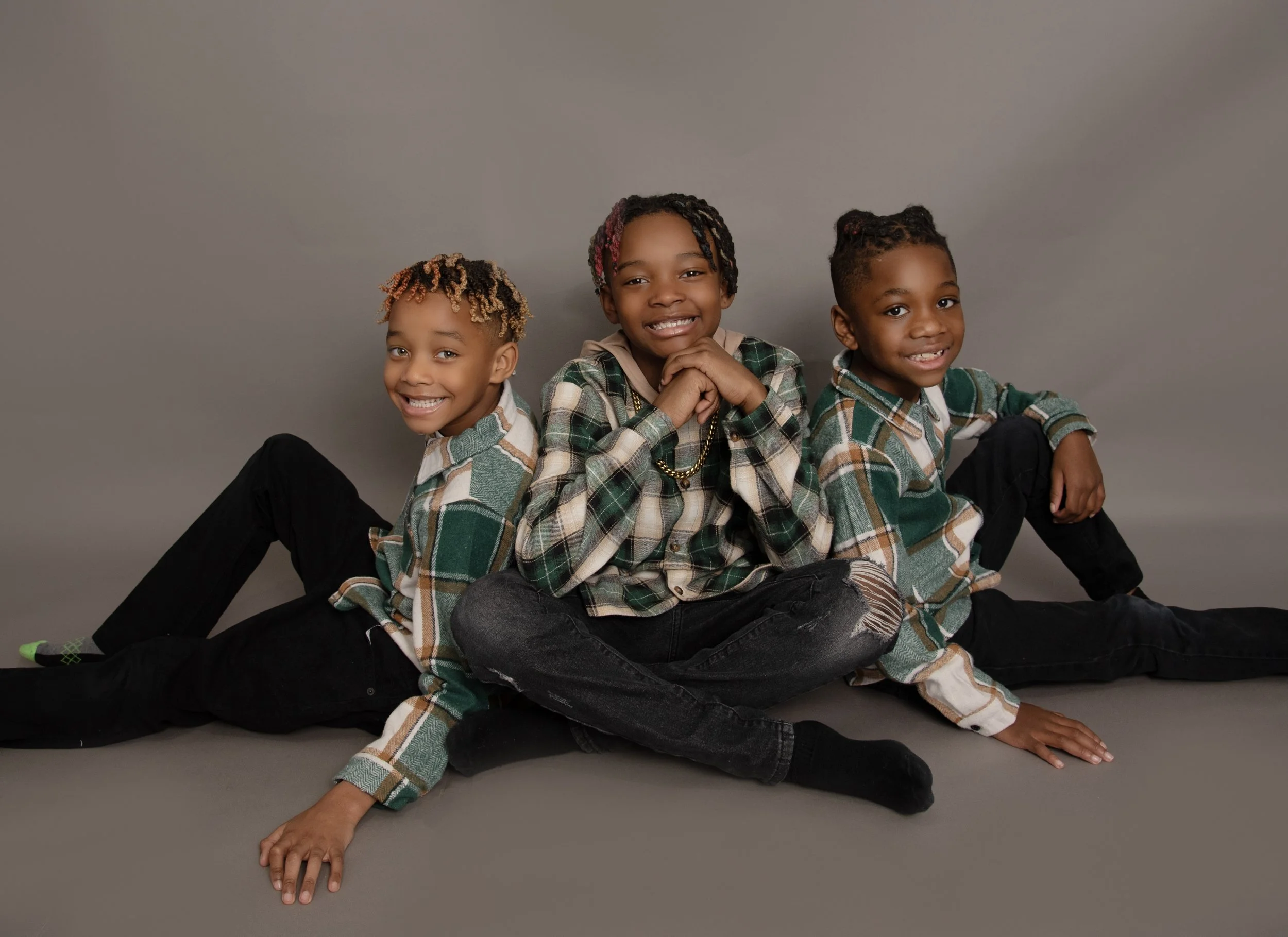 Three children wearing plaid shirts sitting on the floor in front of a gray background.