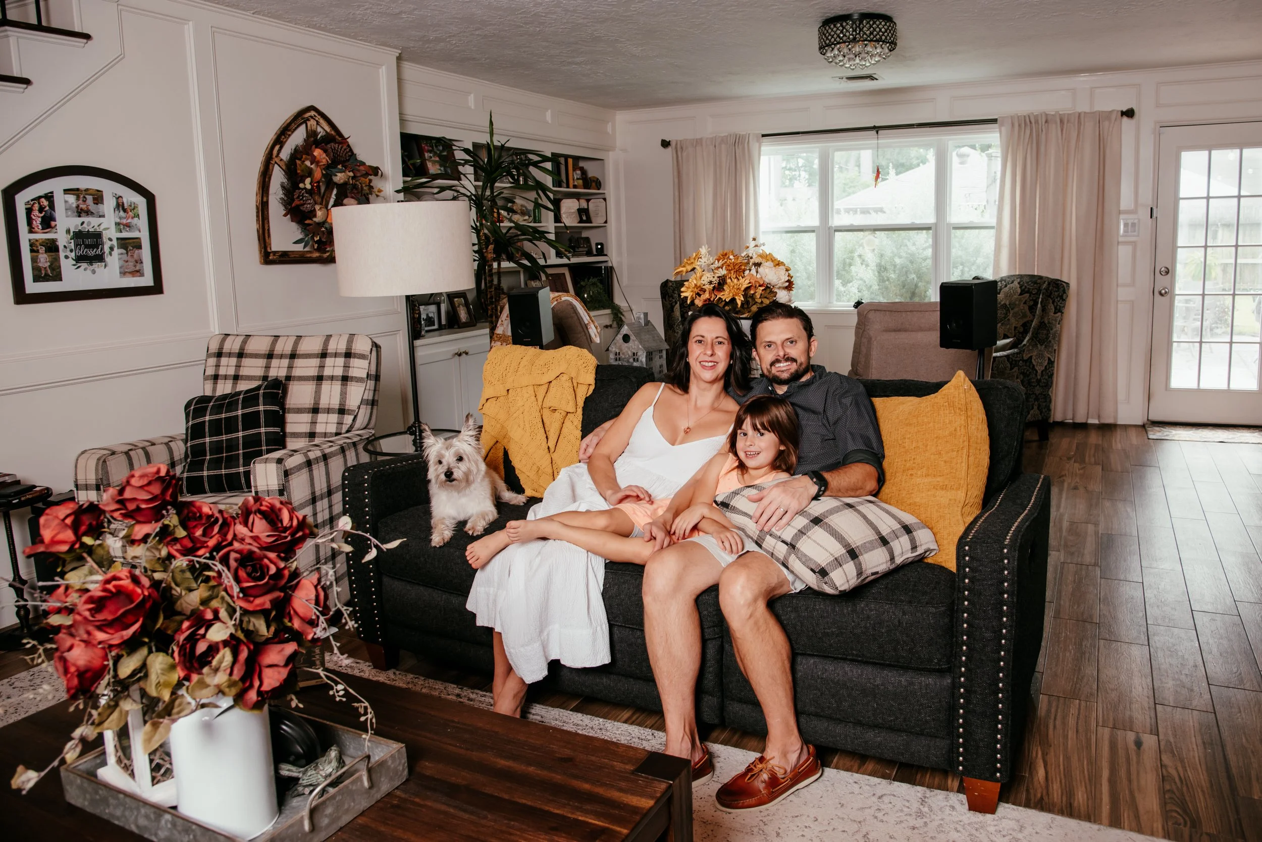 A happy family of three, a man, a woman, and a young girl, sitting together on a dark-colored sofa in a cozy living room, with a small dog sitting beside them. The room has a large window, hardwood floors, and decorative pillows on the sofa.