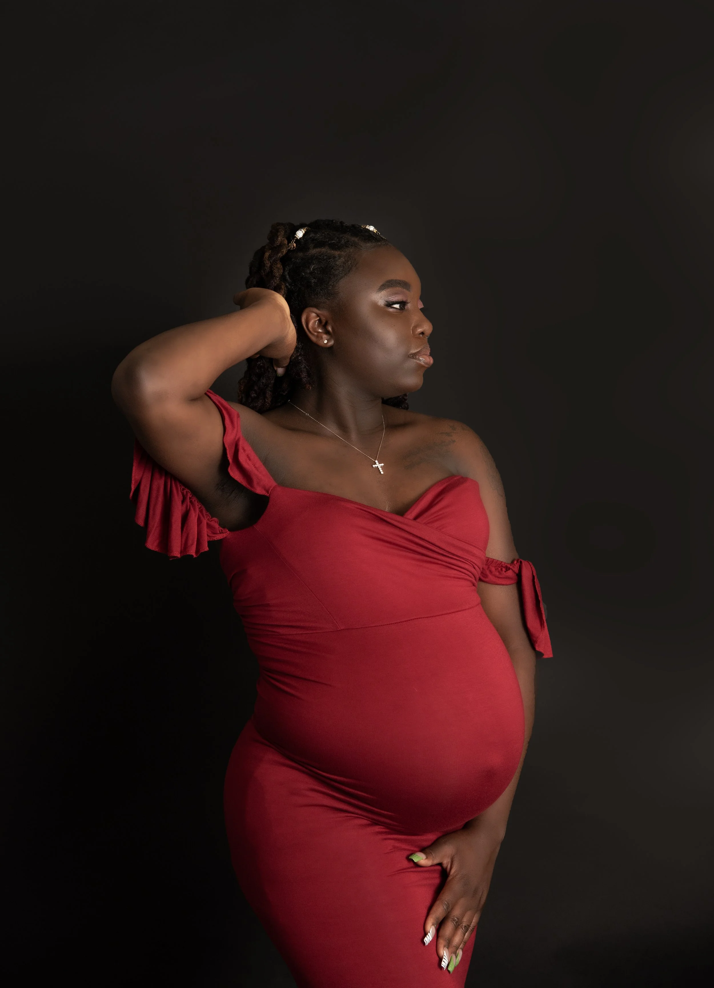 A pregnant woman in a red dress poses with one hand behind her head against a black background.