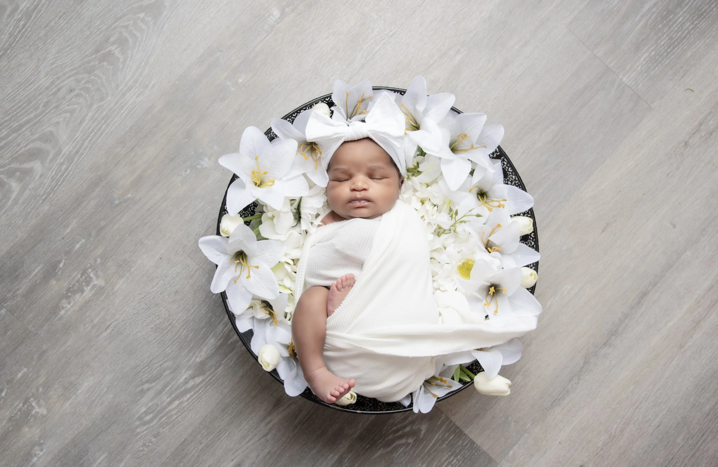 Newborn baby girl wrapped in white laying on bed of lilies in bowl against gray wood floor