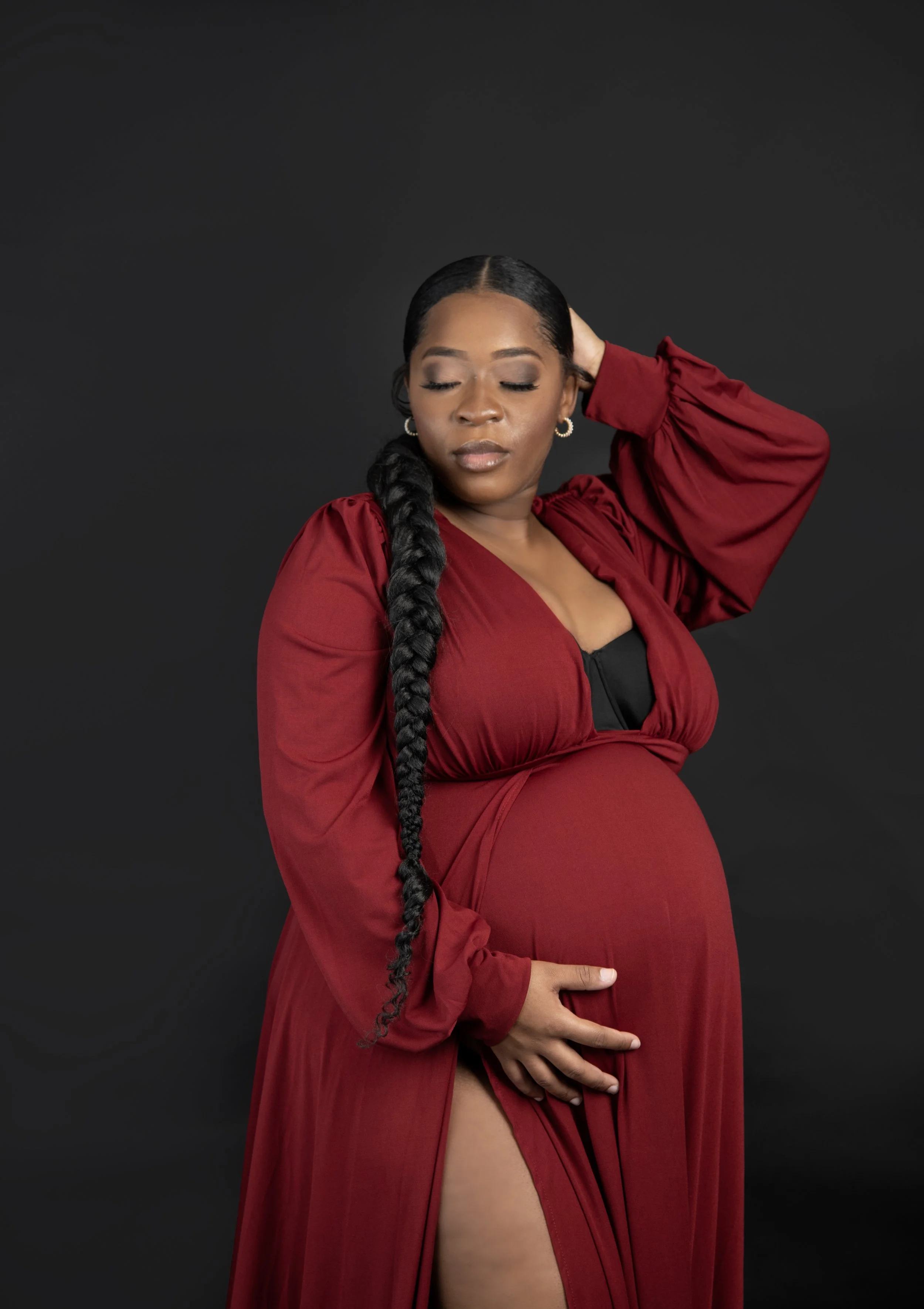 A pregnant woman in a red dress with a slit, posing with her eyes closed against a black background.