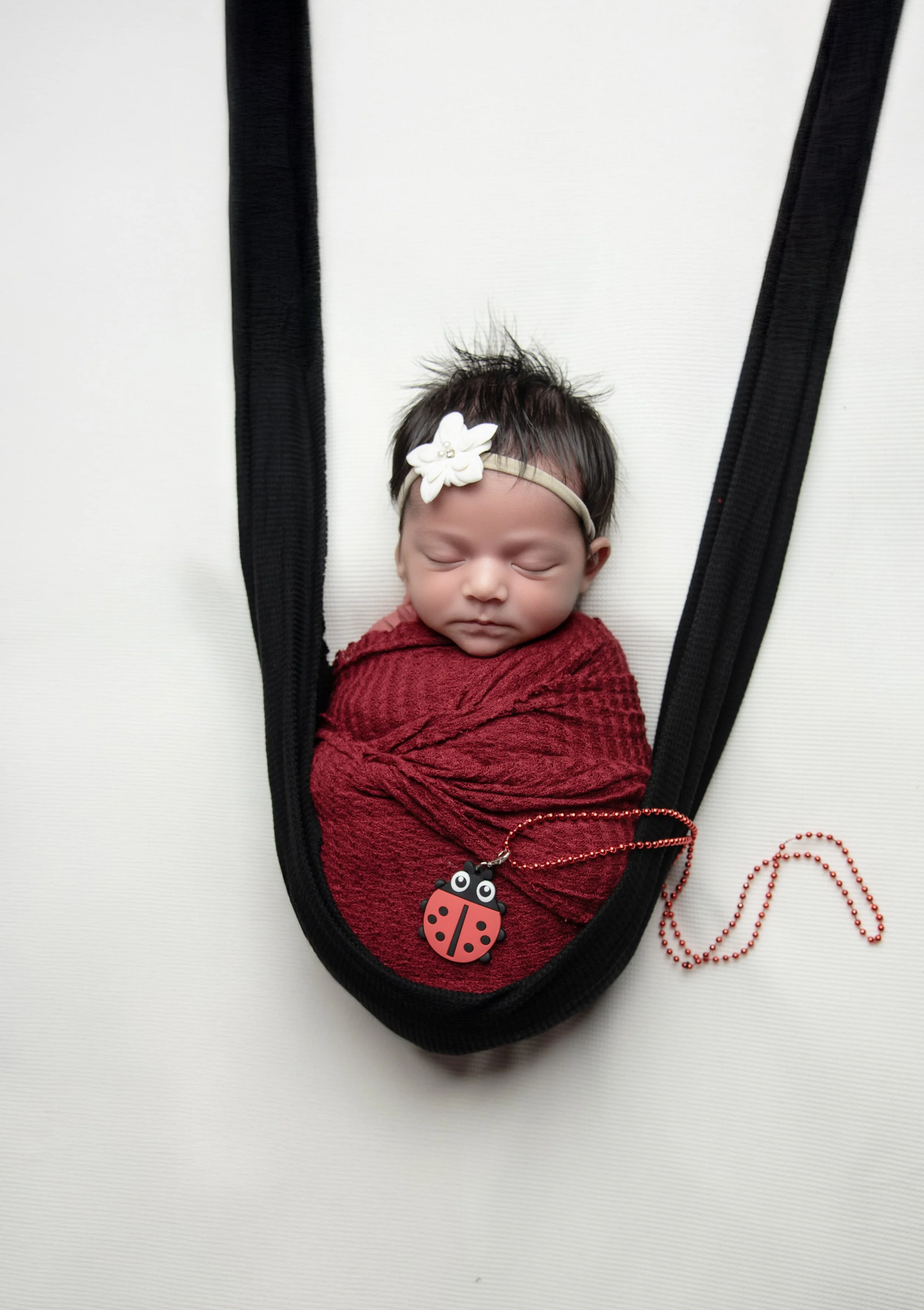 A newborn baby girl sleeping, wrapped in a red blanket, with a white flower headband and a ladybug necklace, lying on a black fabric sling on a white surface.