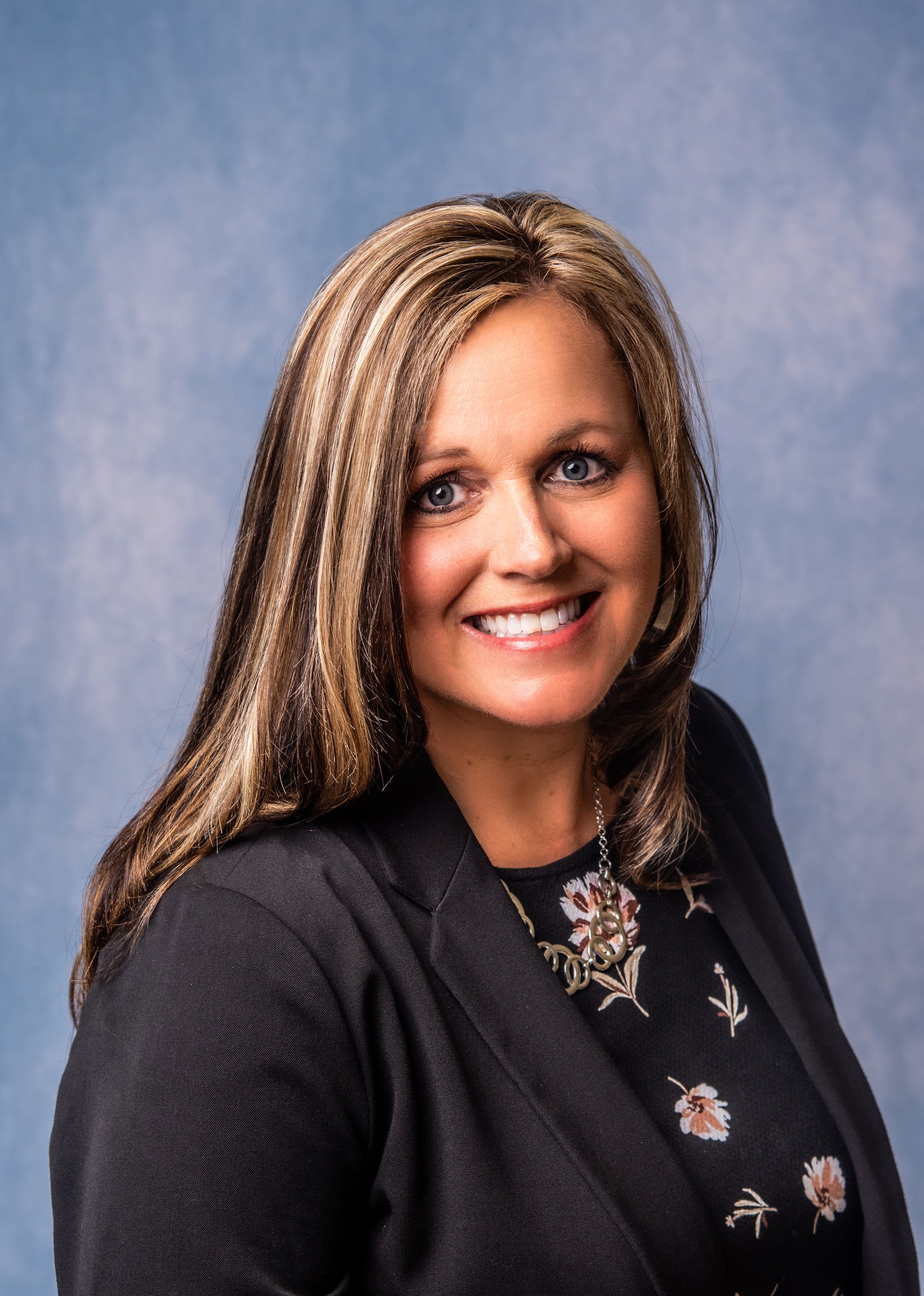 Headshot of a woman with long, brown hair, smiling, wearing a black blazer and floral blouse, with a necklace, against a blue background.