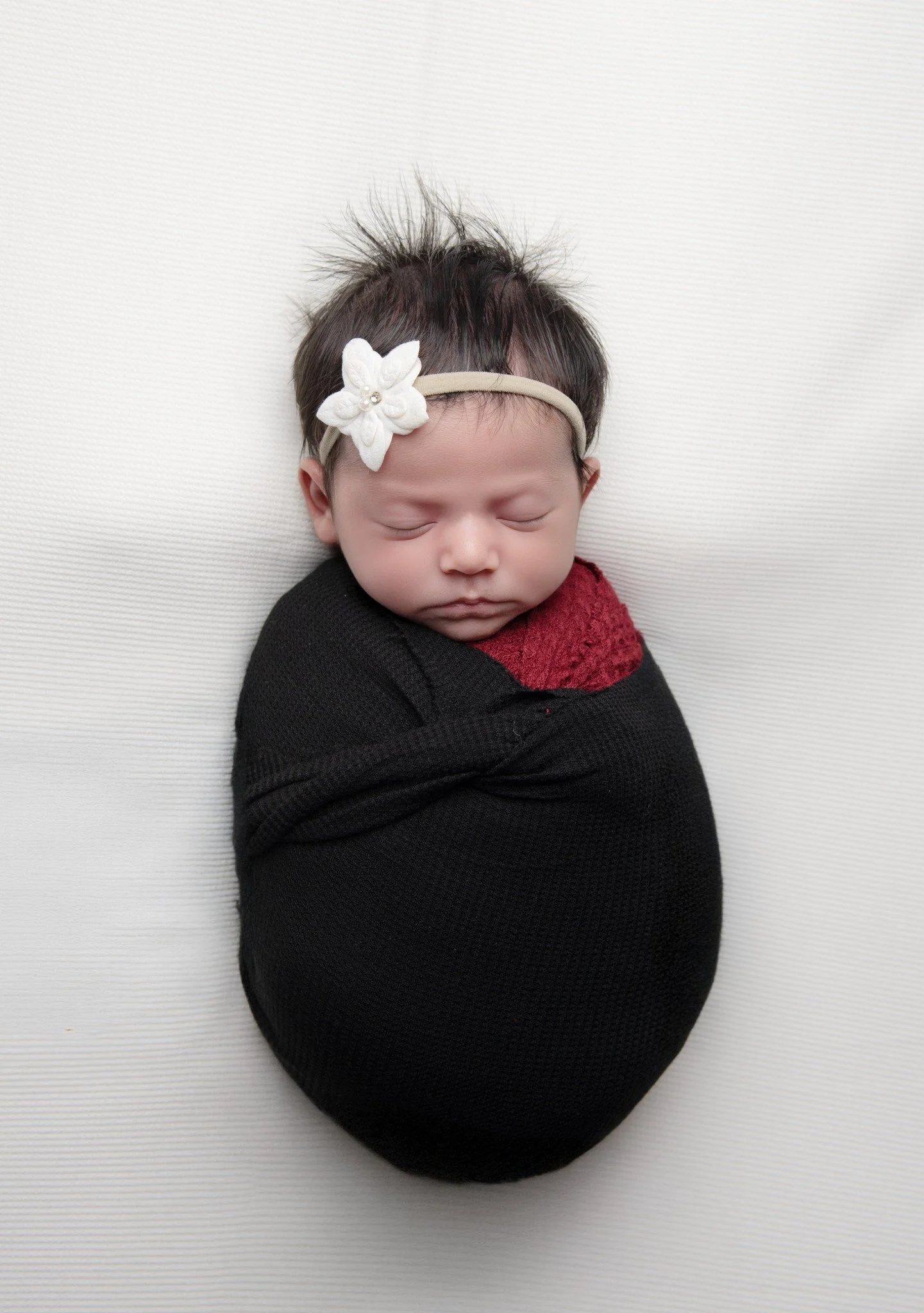 A sleeping newborn baby wrapped in a black swaddle, wearing a burgundy shirt, and a beige headband with a white flower.