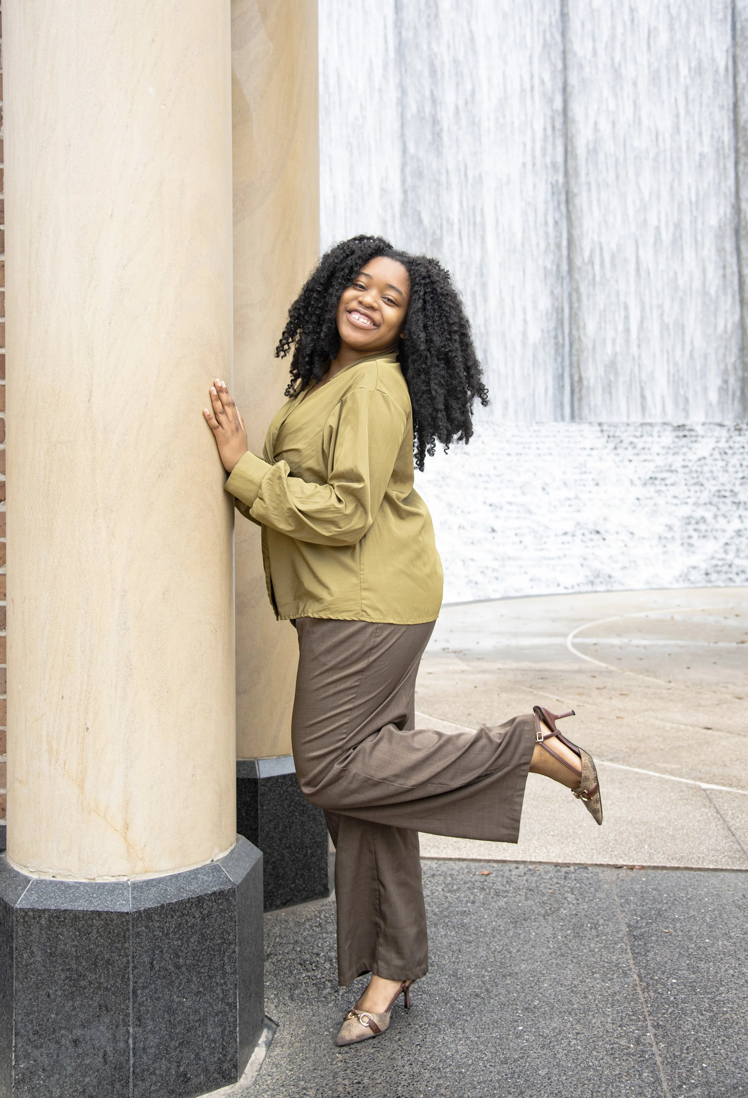 A woman with curly black hair, smiling, wearing a green jacket and brown pants, leaning against a beige column near a wall with a waterfall feature in the background.