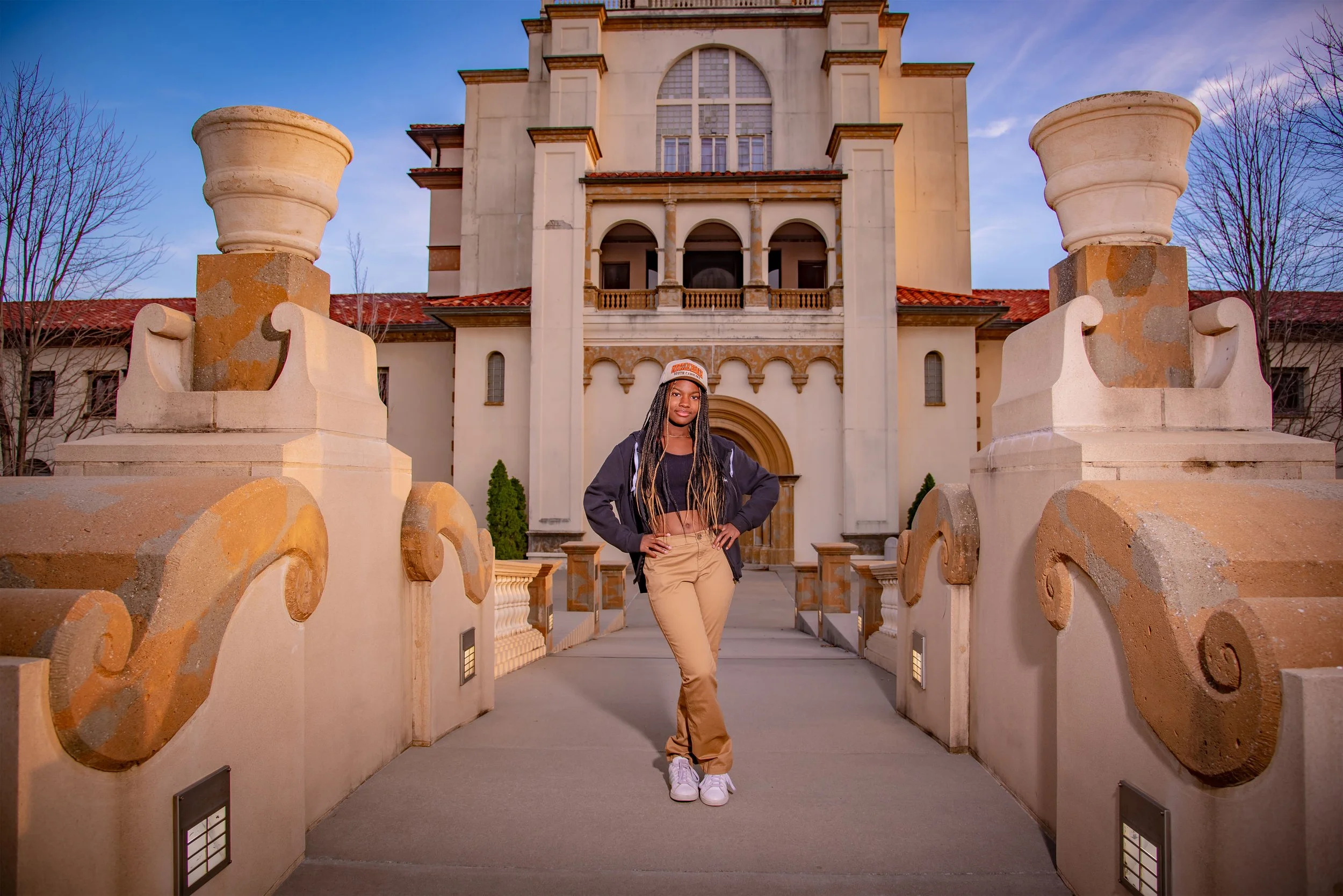 A young woman standing on a walkway in front of a castle-like building, wearing a cap, black jacket, black crop top, tan pants, and white sneakers.