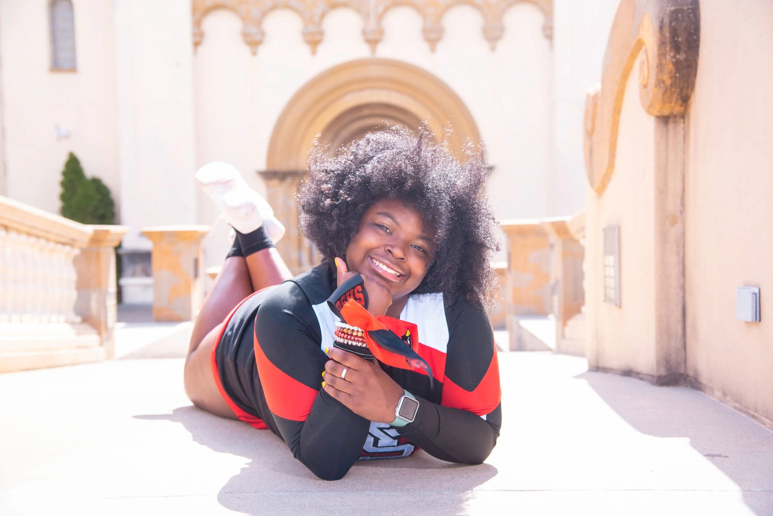 Young woman with curly hair lying on the ground outdoors, smiling, wearing sportswear, with a sunny background featuring a building with arches.