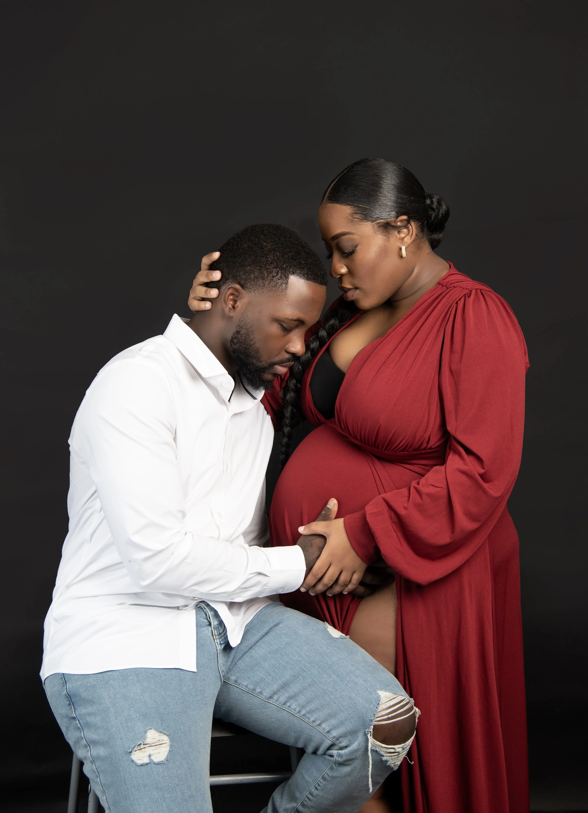 A pregnant woman in a red dress and her partner are holding hands and sharing a moment of affection against a black studio backdrop.