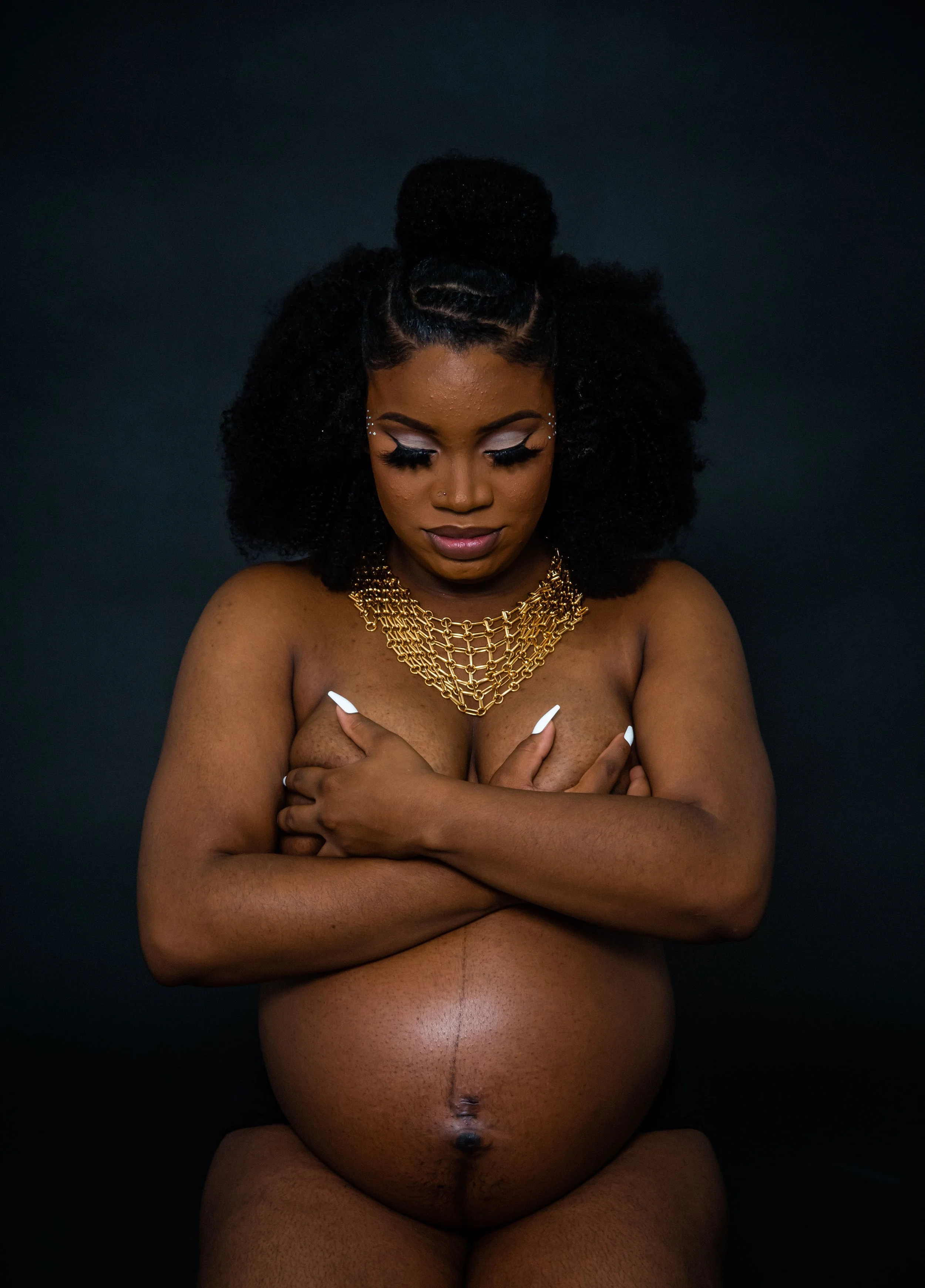 A pregnant woman with dark skin and curly black hair, styled with a high bun, standing against a dark background. She is topless, with her arms crossed over her chest, and is wearing a layered gold necklace. She has eye makeup with silver eyeshadow a