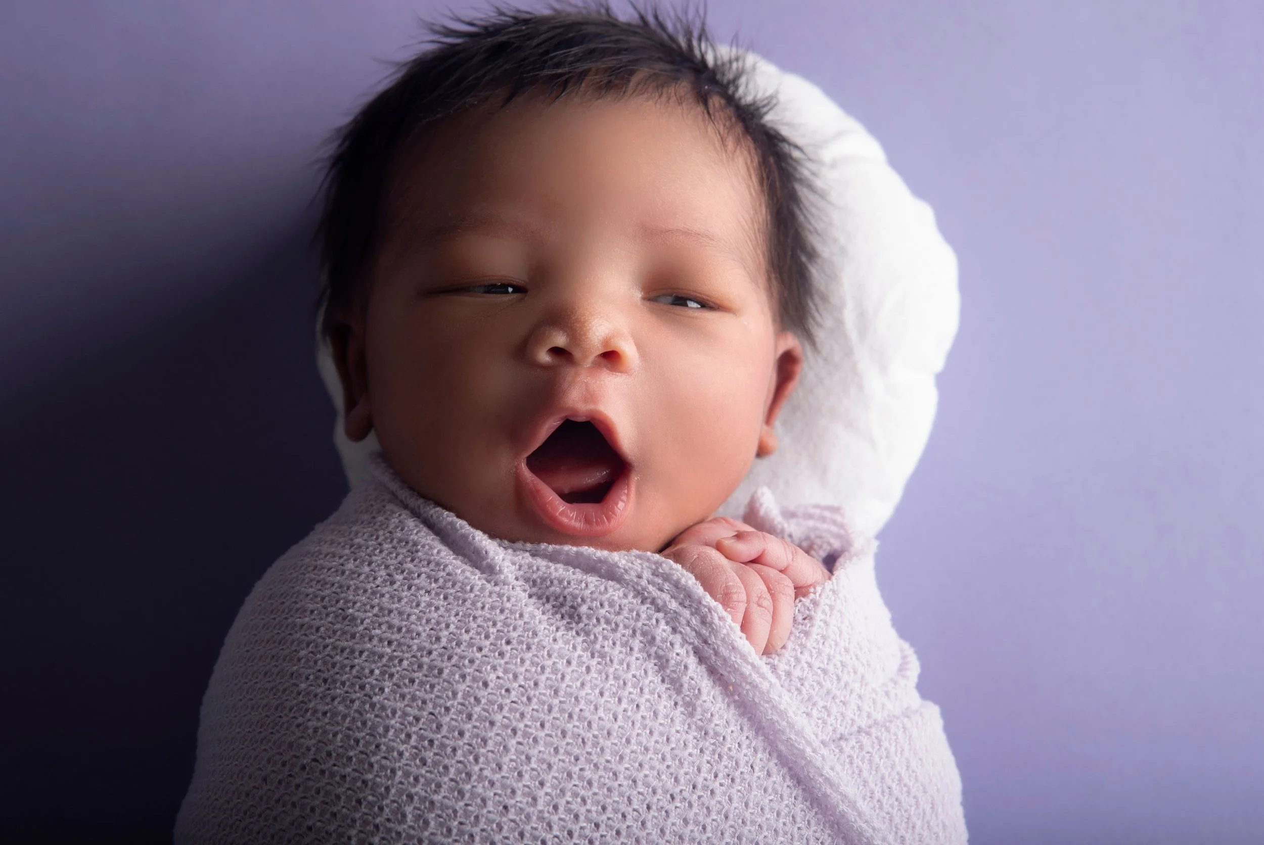 A yawning infant wrapped in a light pink blanket, lying on a white pillow with a purple background.