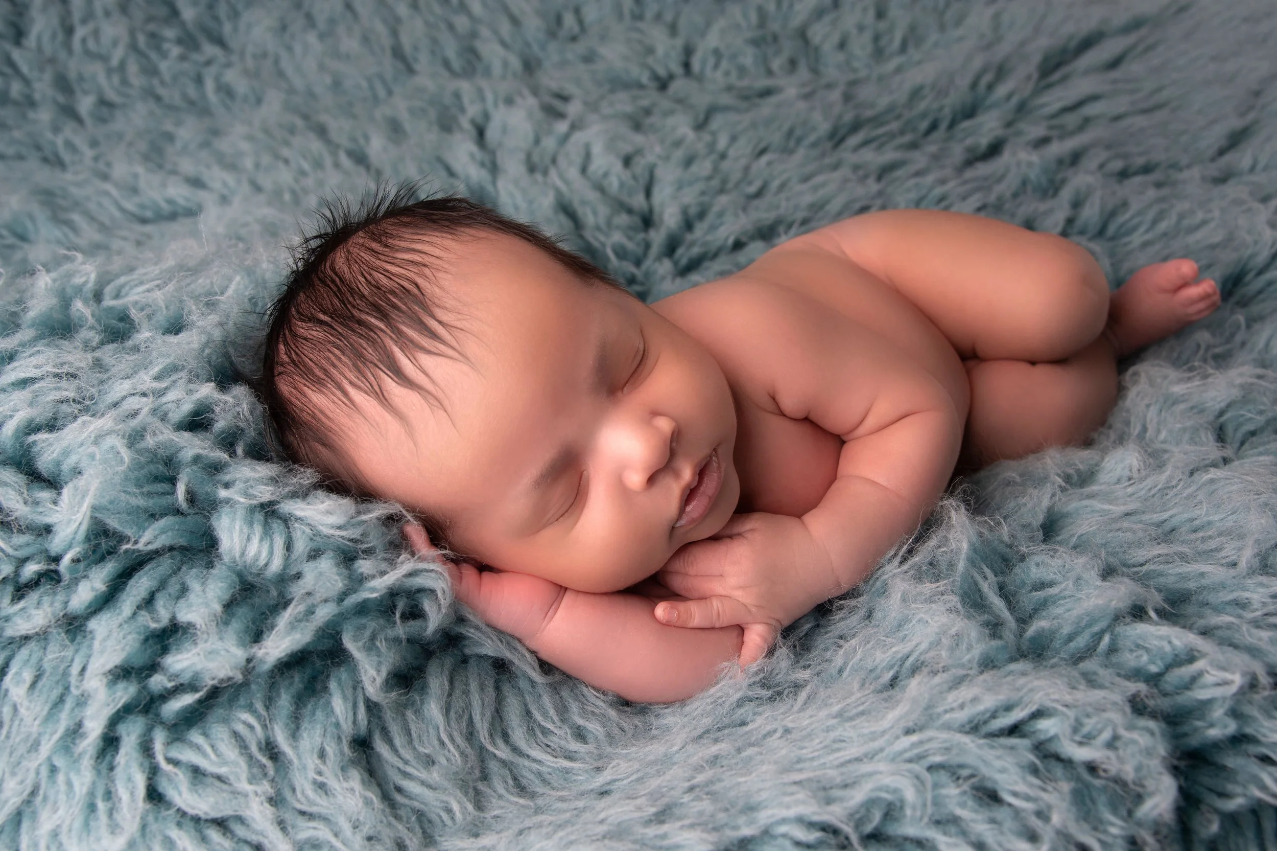 A sleeping newborn baby lying on a soft, textured blue blanket with their hands tucked under their head.
