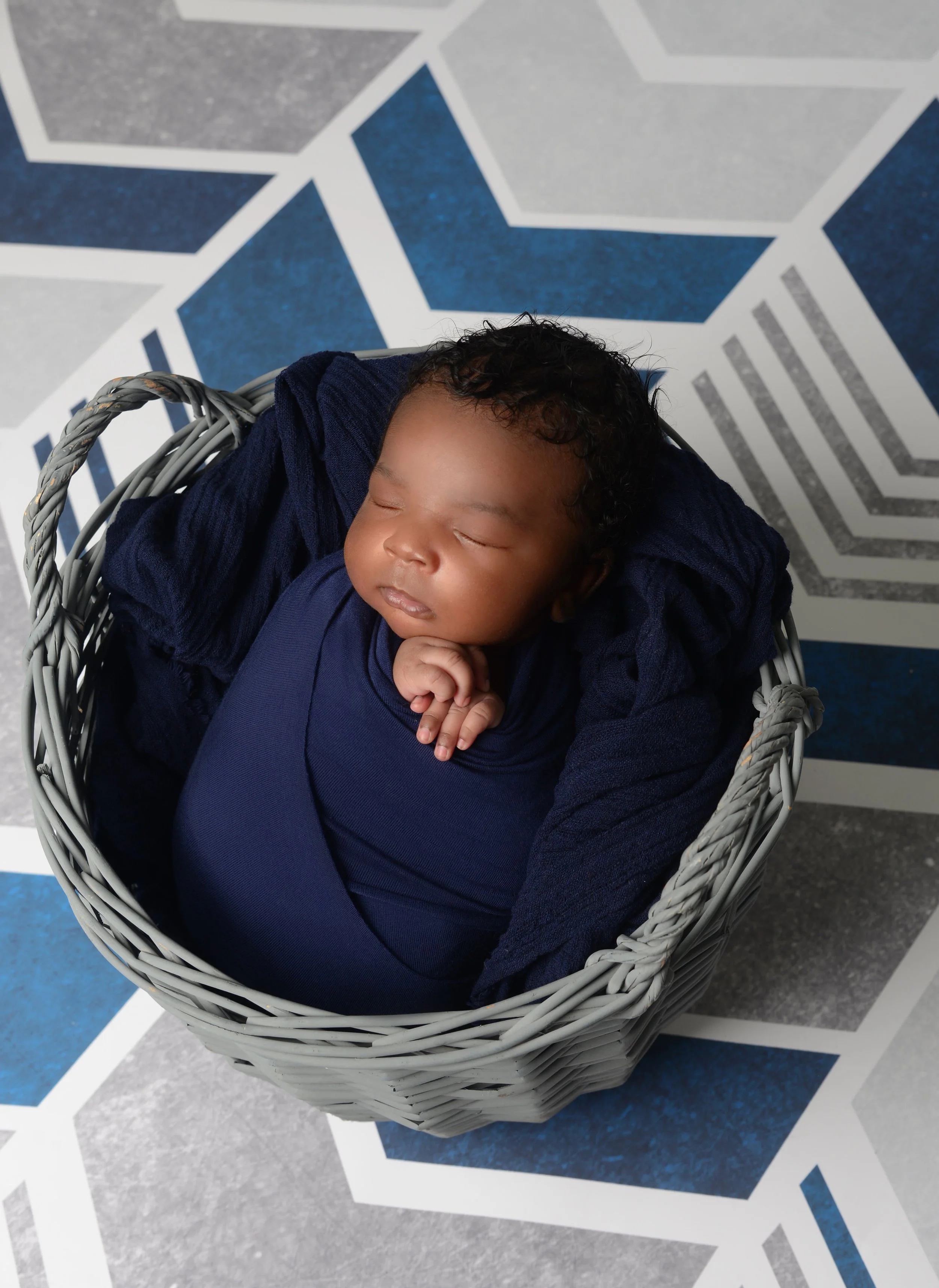 A sleeping baby wrapped in navy blue fabric, lying in a gray wicker basket lined with a dark blue blanket, on a geometric patterned floor in shades of gray, white, and dark blue.