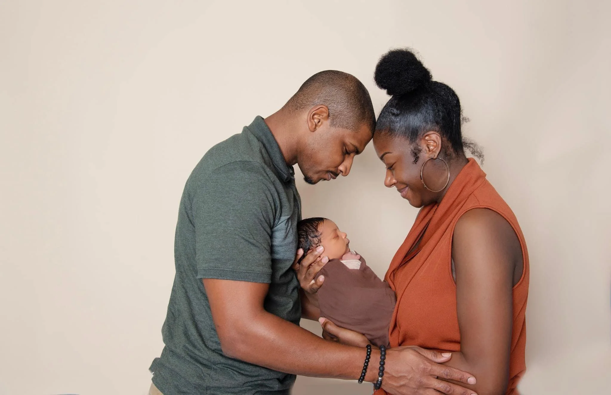 Mom and dad head to head holding newborn baby boy wrapped in brown fabric against beige background