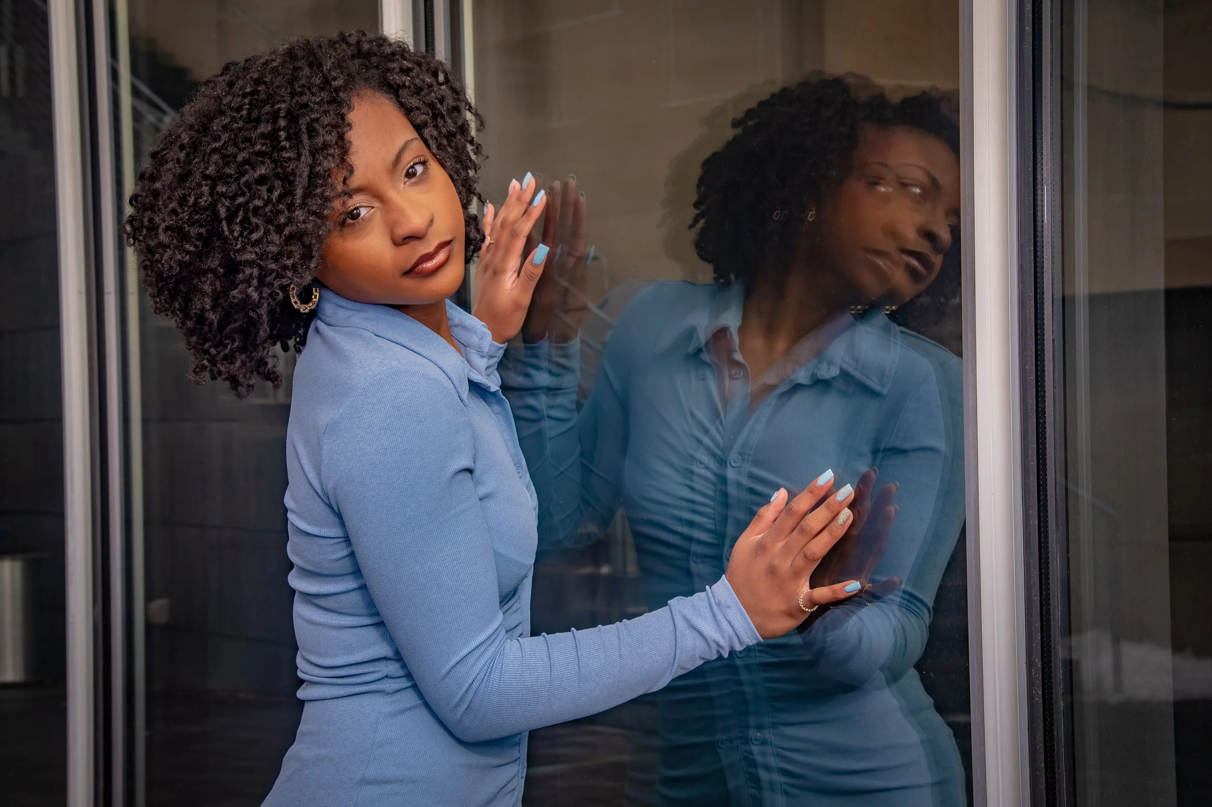 A woman with curly hair and wearing a blue shirt stands beside a glass door, with her hands on the glass, looking at the camera, as a reflection of her face appears in the glass.