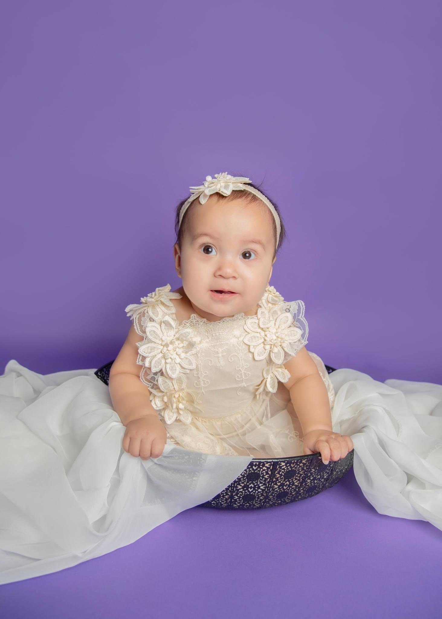 Adorable baby girl wearing a cream-colored dress with lace and floral details, sitting in a decorative black basket on a purple background, with a matching headband.