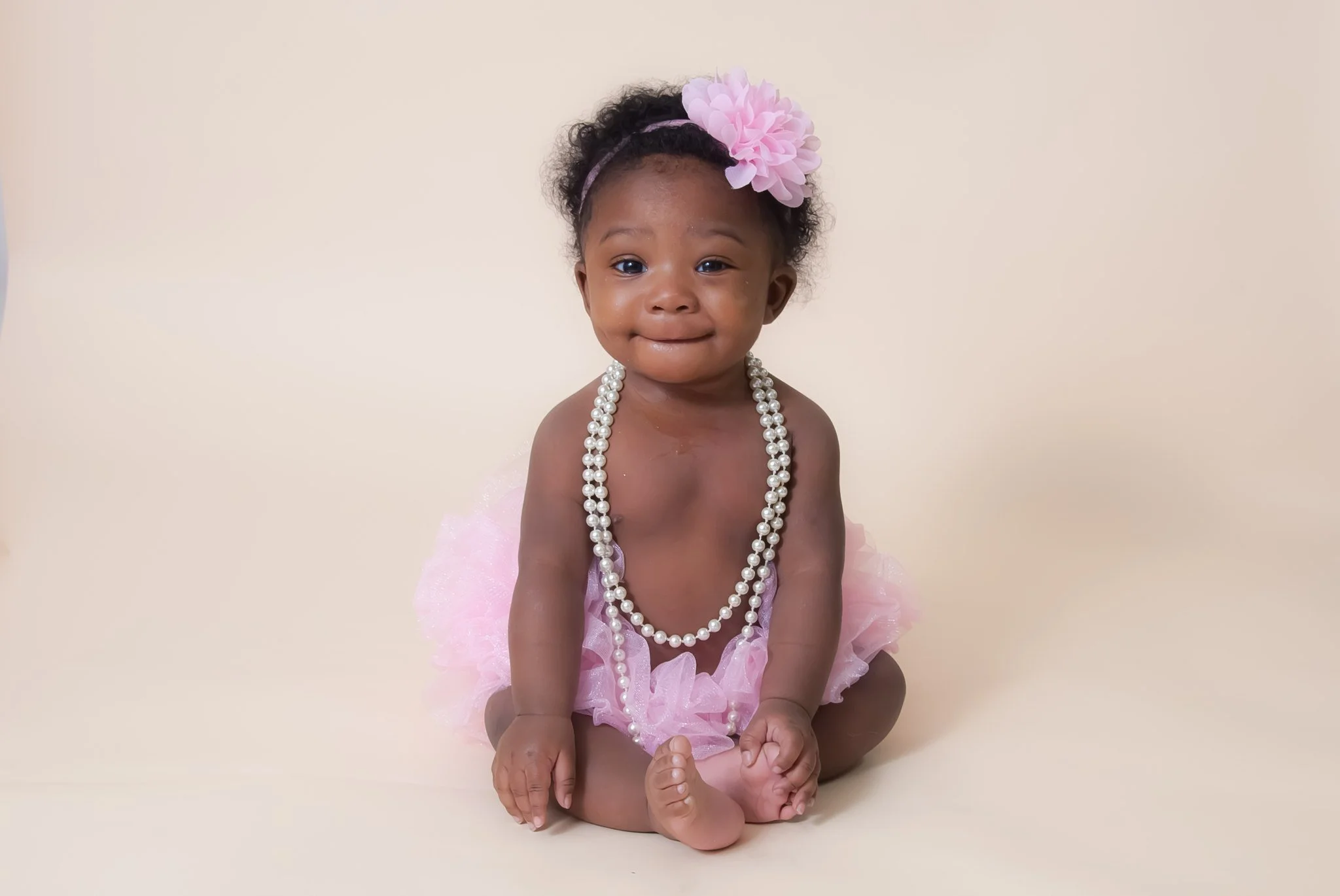 Adorable baby girl sitting on a plain beige background, wearing a pink tutu, a headband with a large pink flower, and a long string of faux pearls, smiling softly.