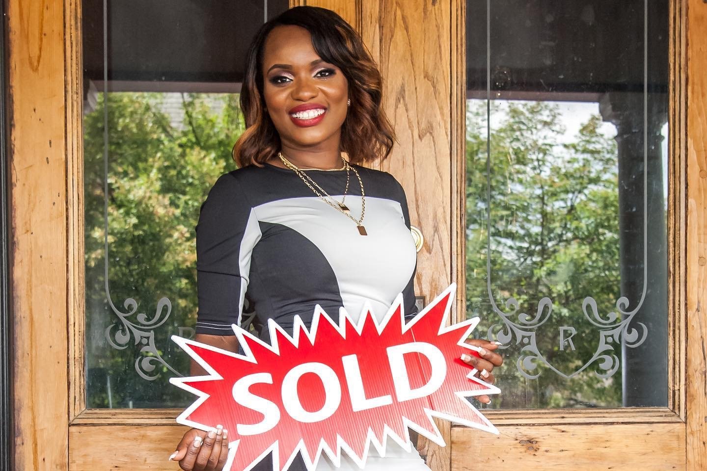 A realtor with shoulder-length brown hair, smiling, holding a red and white 'SOLD' sign in front of a wooden door with glass windows.