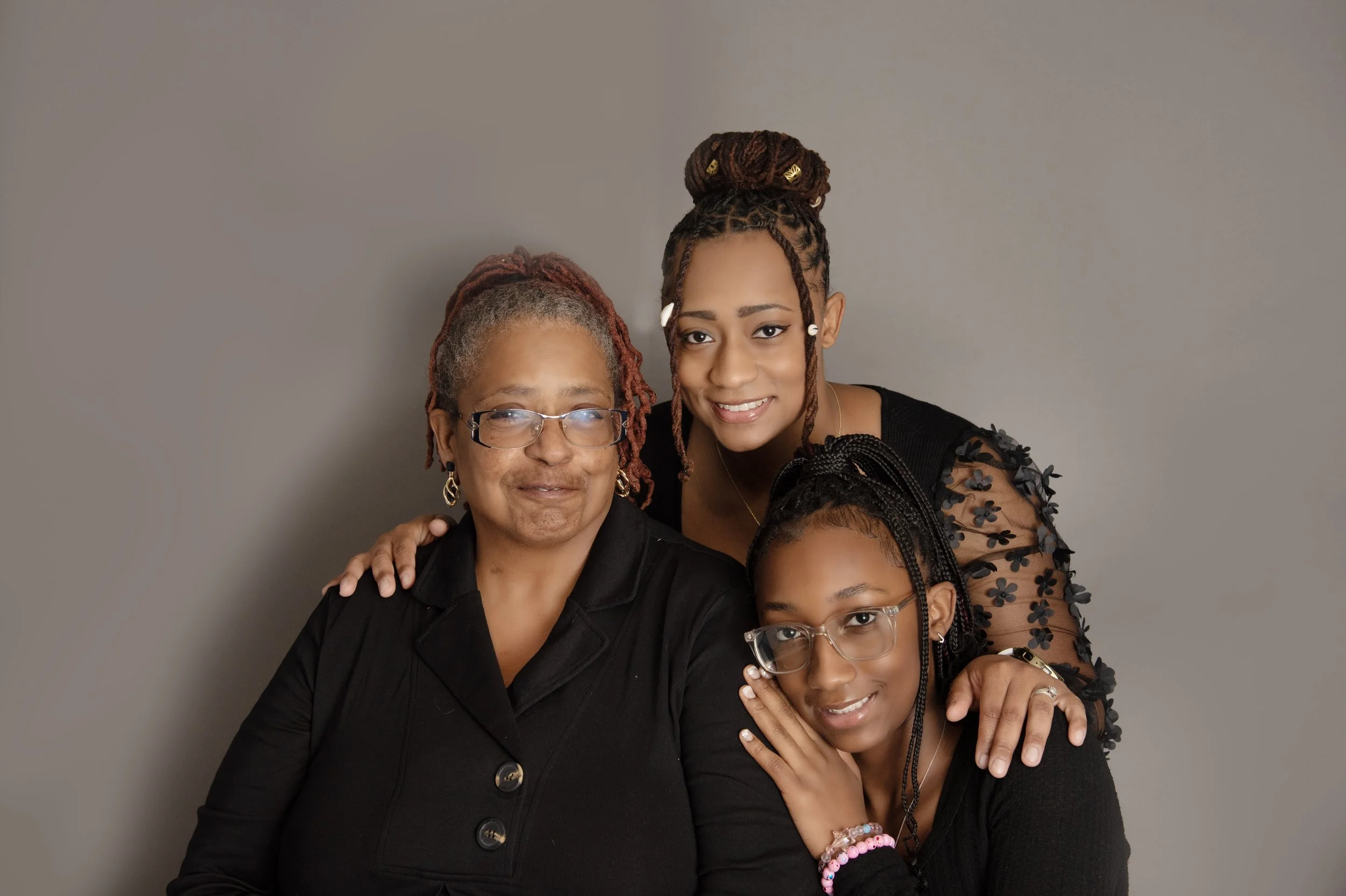 Three women, possibly a family, posing together against a plain gray background. The older woman on the left has gray dreadlocks, glasses, and is wearing a black top. The young woman in the middle has styled dreadlocks in a bun with gold accents and 