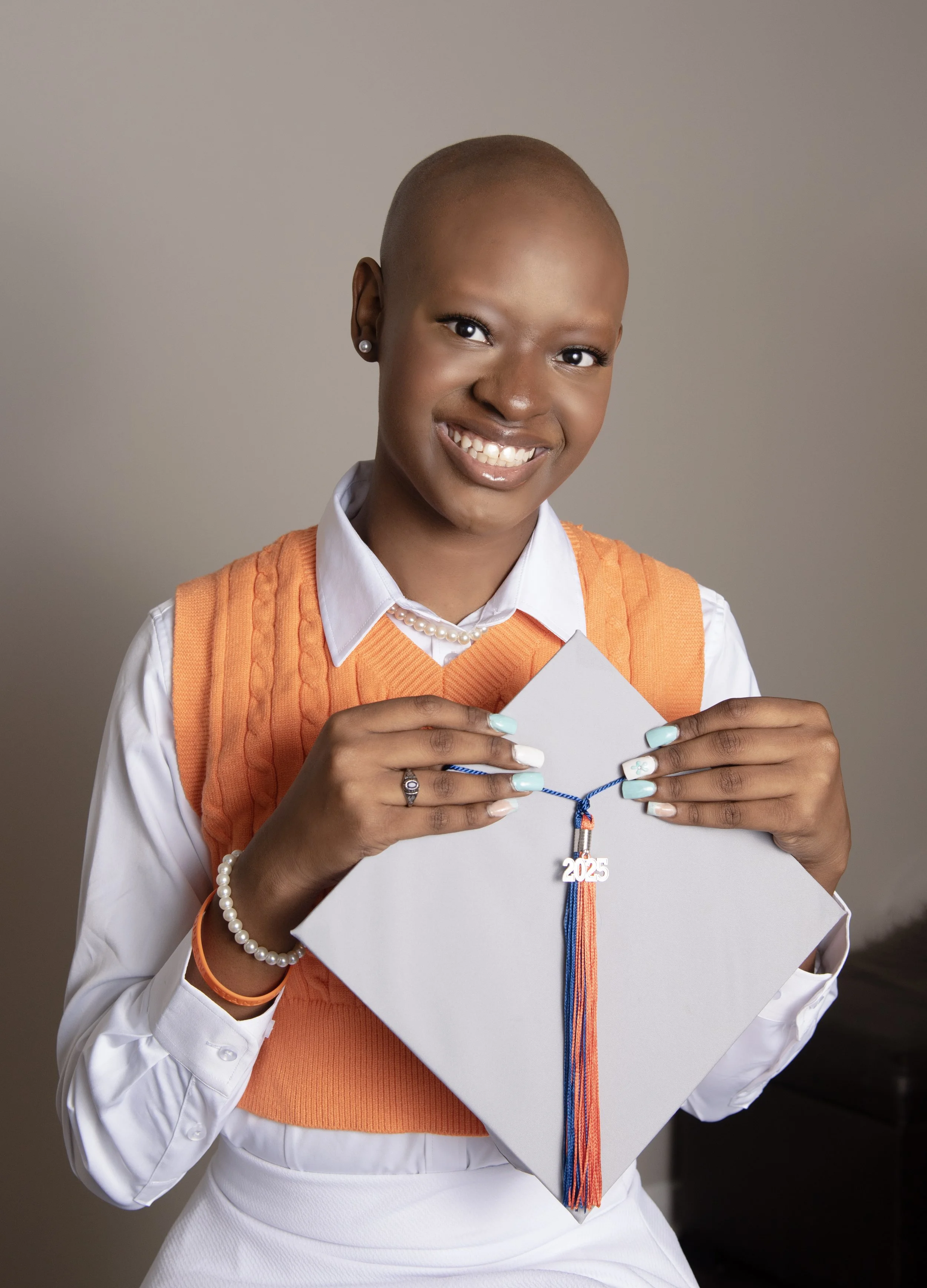 Woman with a shaved head smiling, holding a white cake with a blue and orange tassel necklace and a '2025' medal, wearing an orange sweater vest over a white shirt.