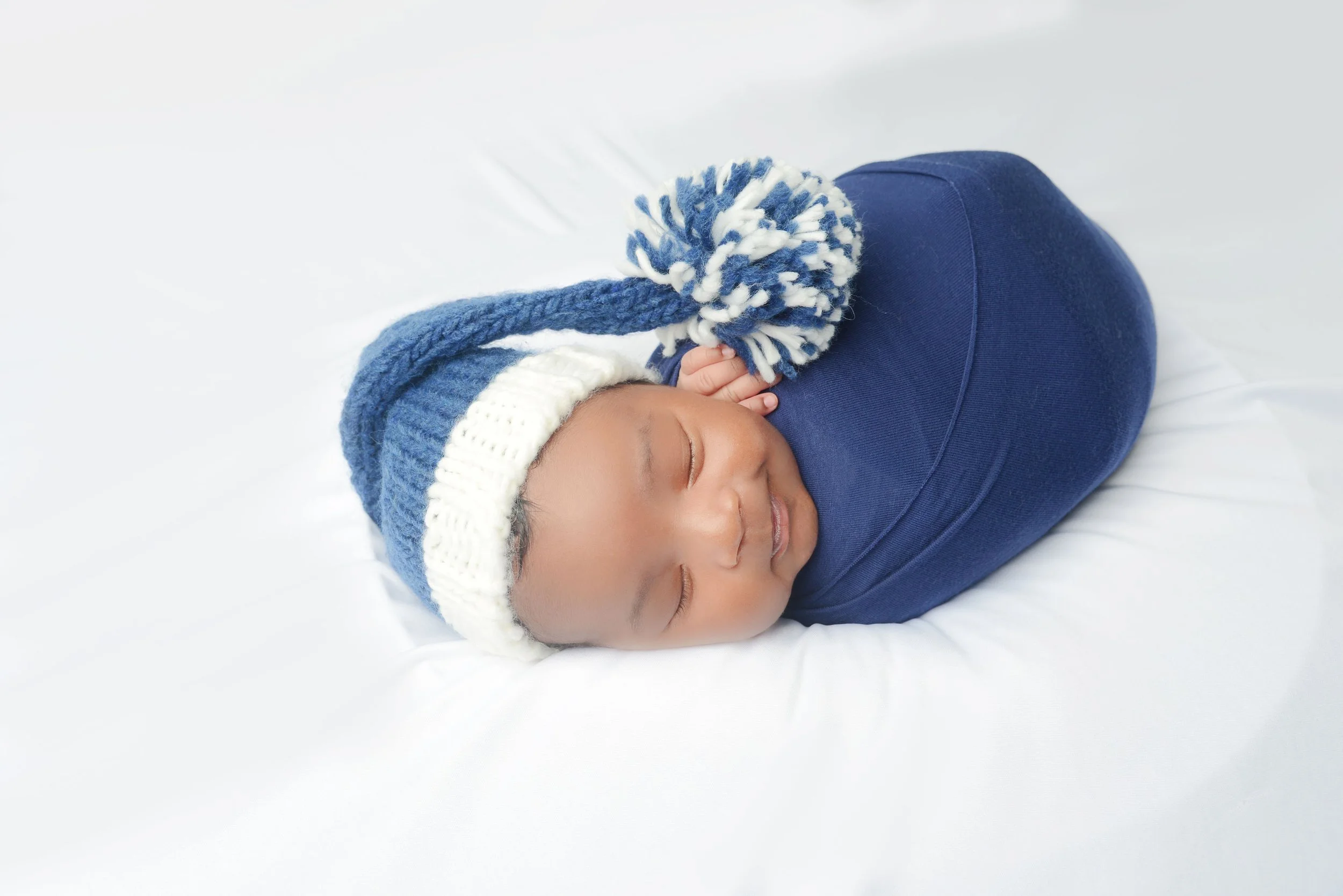 A sleeping baby wearing a blue and white knit hat with a pom-pom and a matching blue outfit, lying on a white surface.