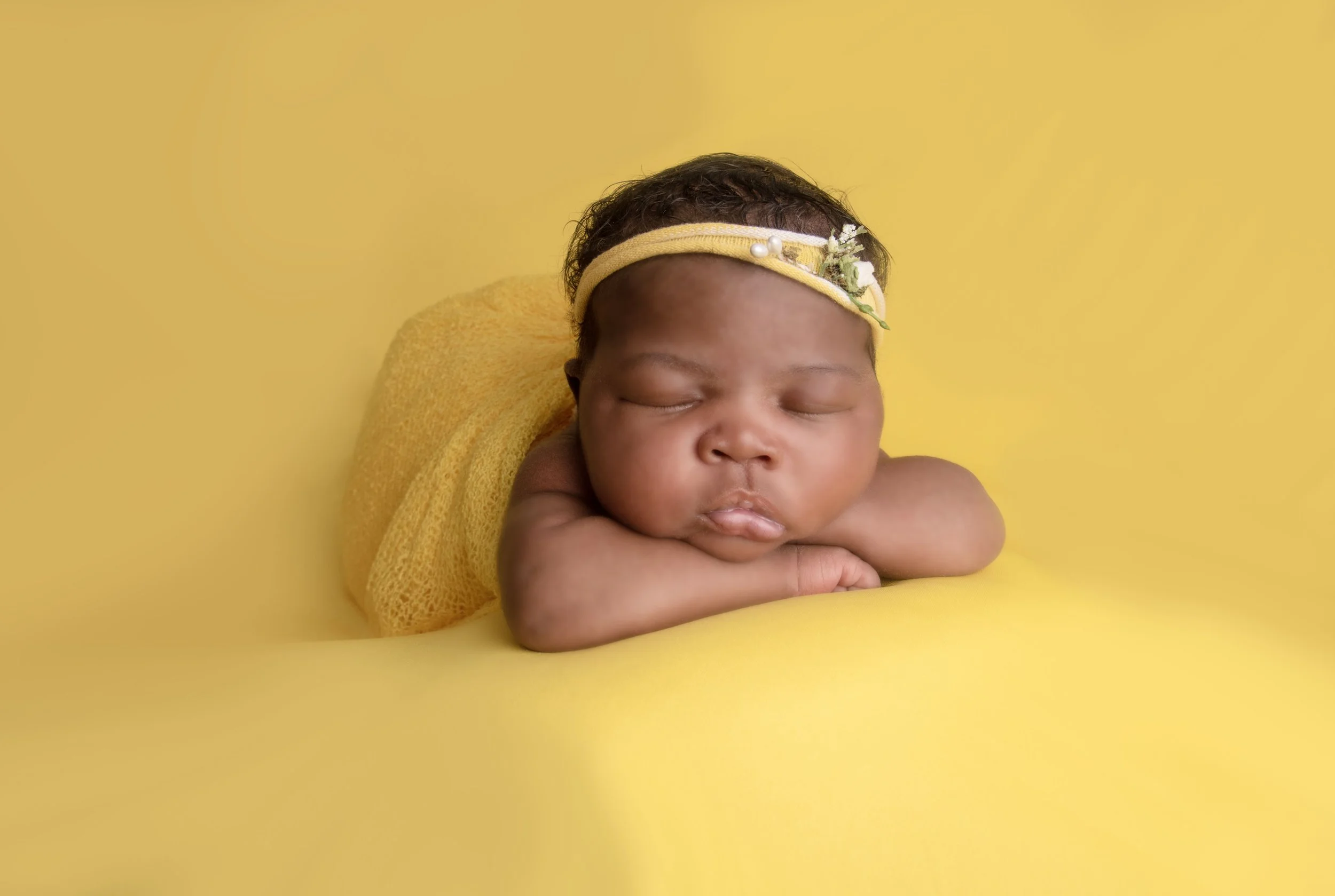 A sleeping baby with a yellow headband, wrapped in yellow cloth, resting against a yellow background.