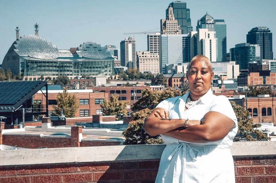 A woman with short hair and arms crossed, standing on a rooftop with a city skyline in the background including modern high-rise buildings and a modern convention center.