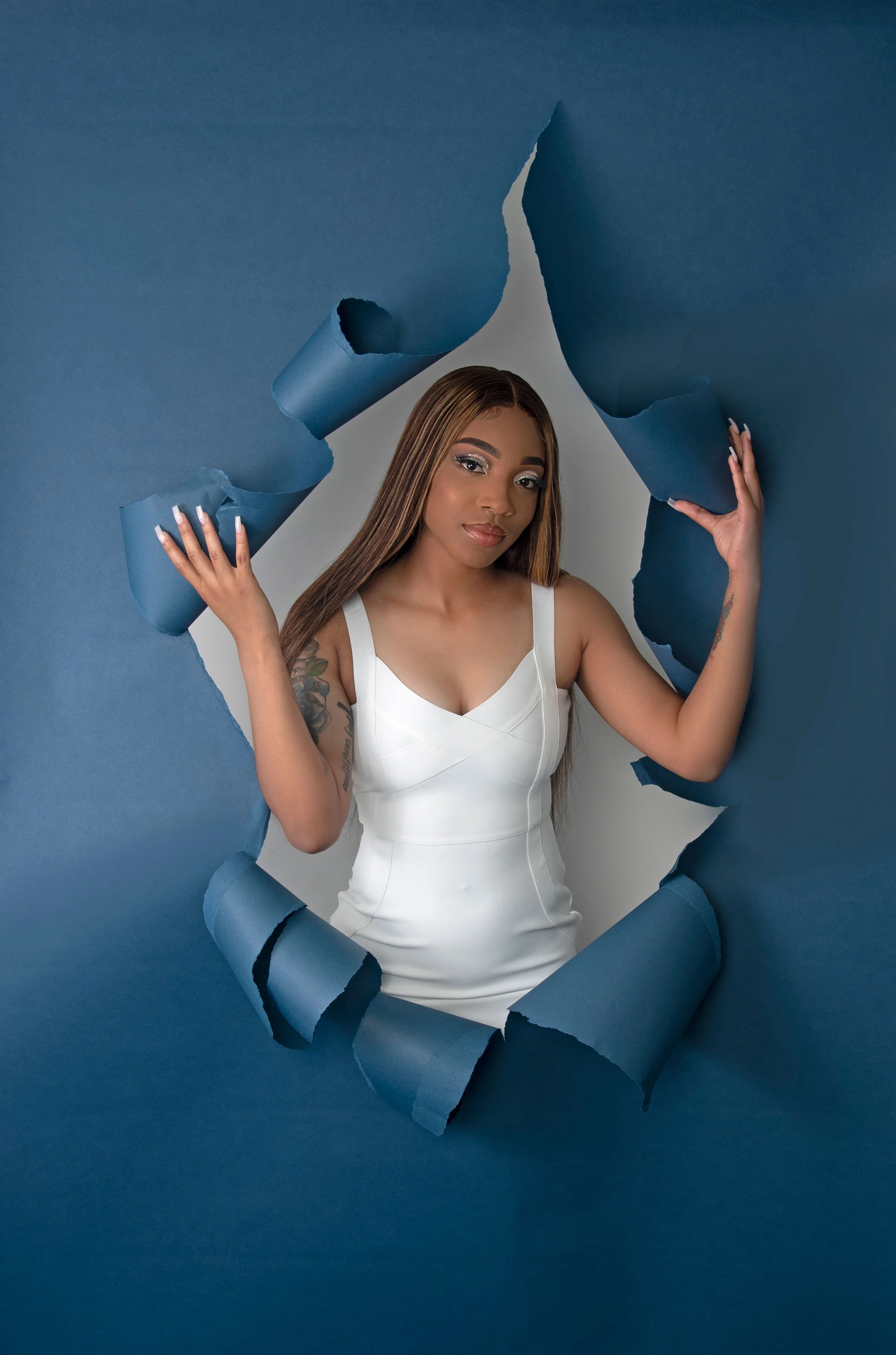 A woman in a white dress is breaking through a blue paper backdrop, holding the torn edges, with her long brown hair and tattooed arm visible.