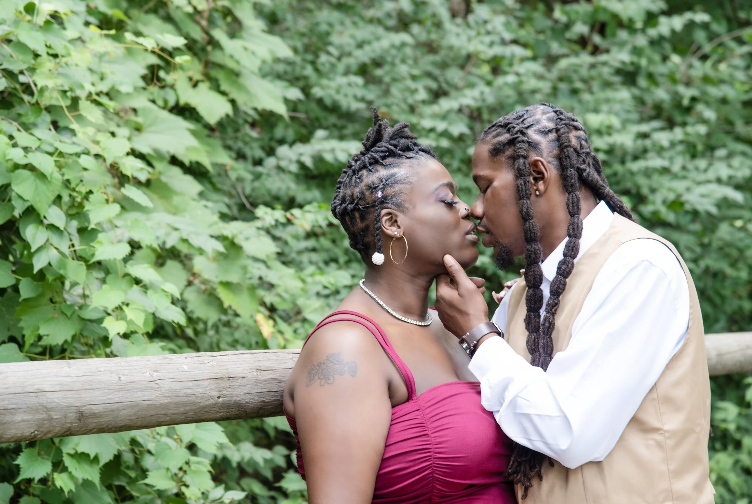 A couple with dreadlocks and earrings embraces tenderly outdoors in front of green foliage.