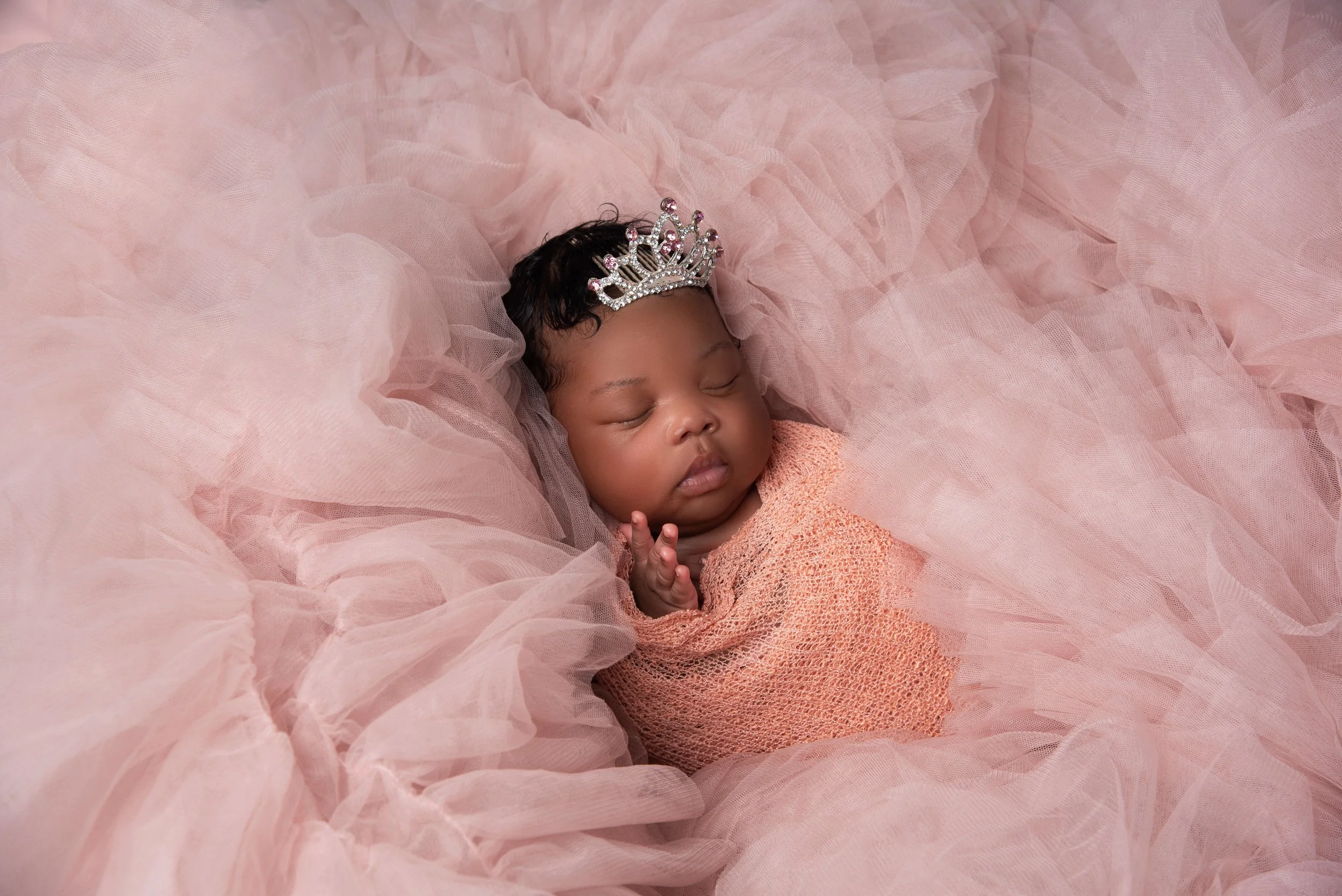 A sleeping baby girl wearing a silver tiara and wrapped in pink tulle fabric.