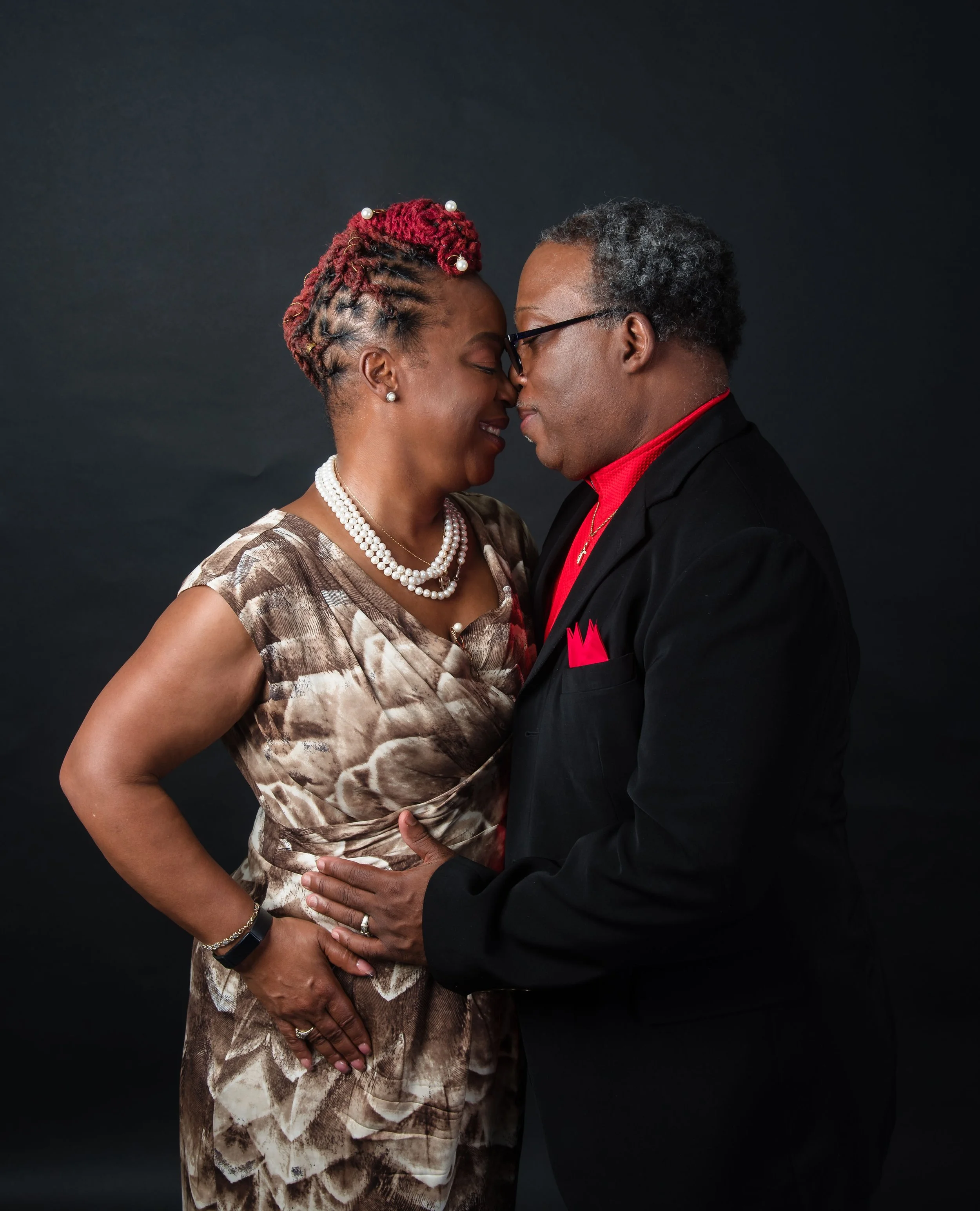 An African American couple touching foreheads with eyes closed, smiling, against a dark background. The woman has short, red-dyed hair styled in twists with pearl accessories, wearing a brown and beige patterned dress with pearl jewelry. The man has 