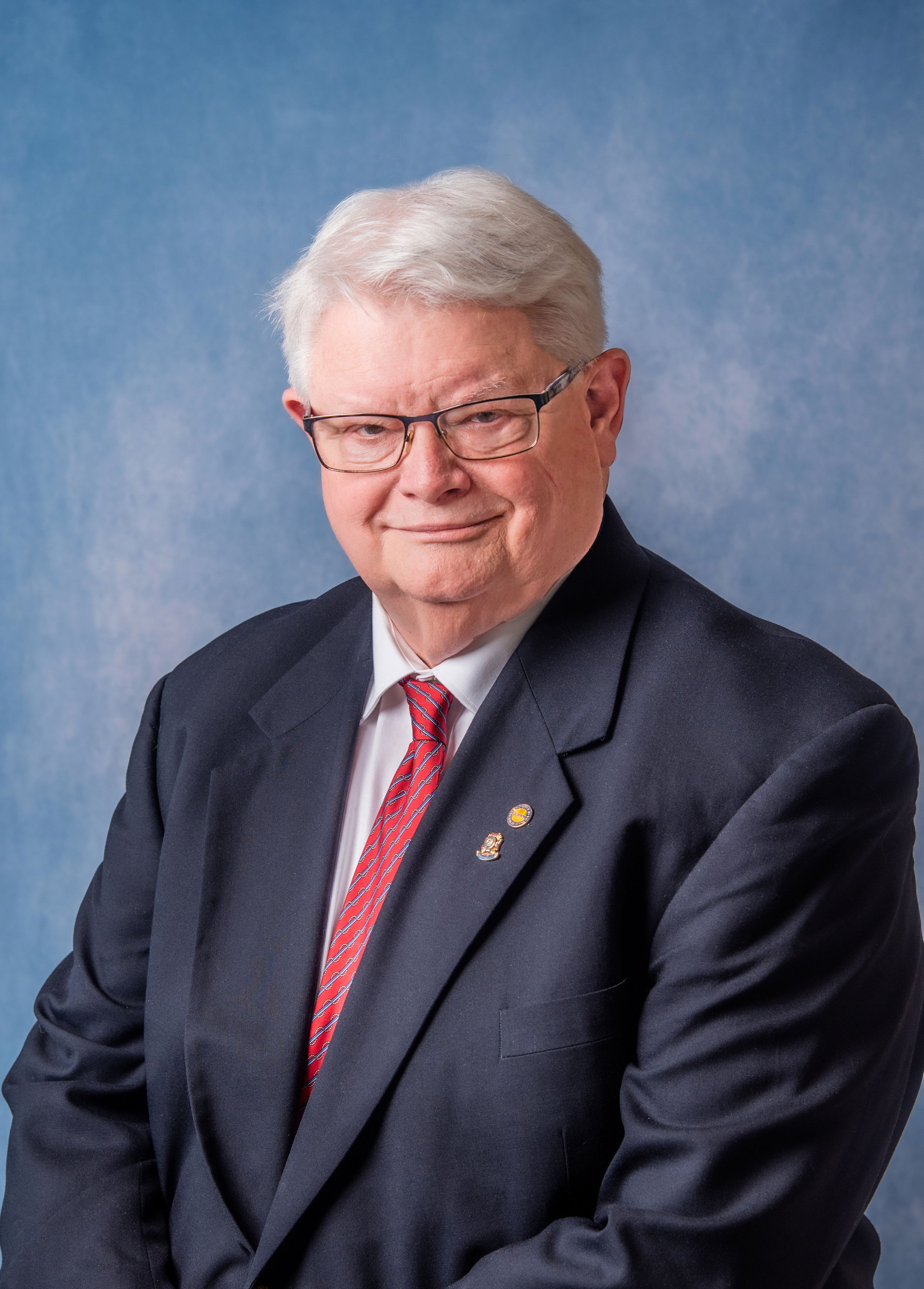 A professional headshot of man with white hair and glasses, wearing a dark suit, white shirt, and red patterned tie, smiling slightly against a blue background.