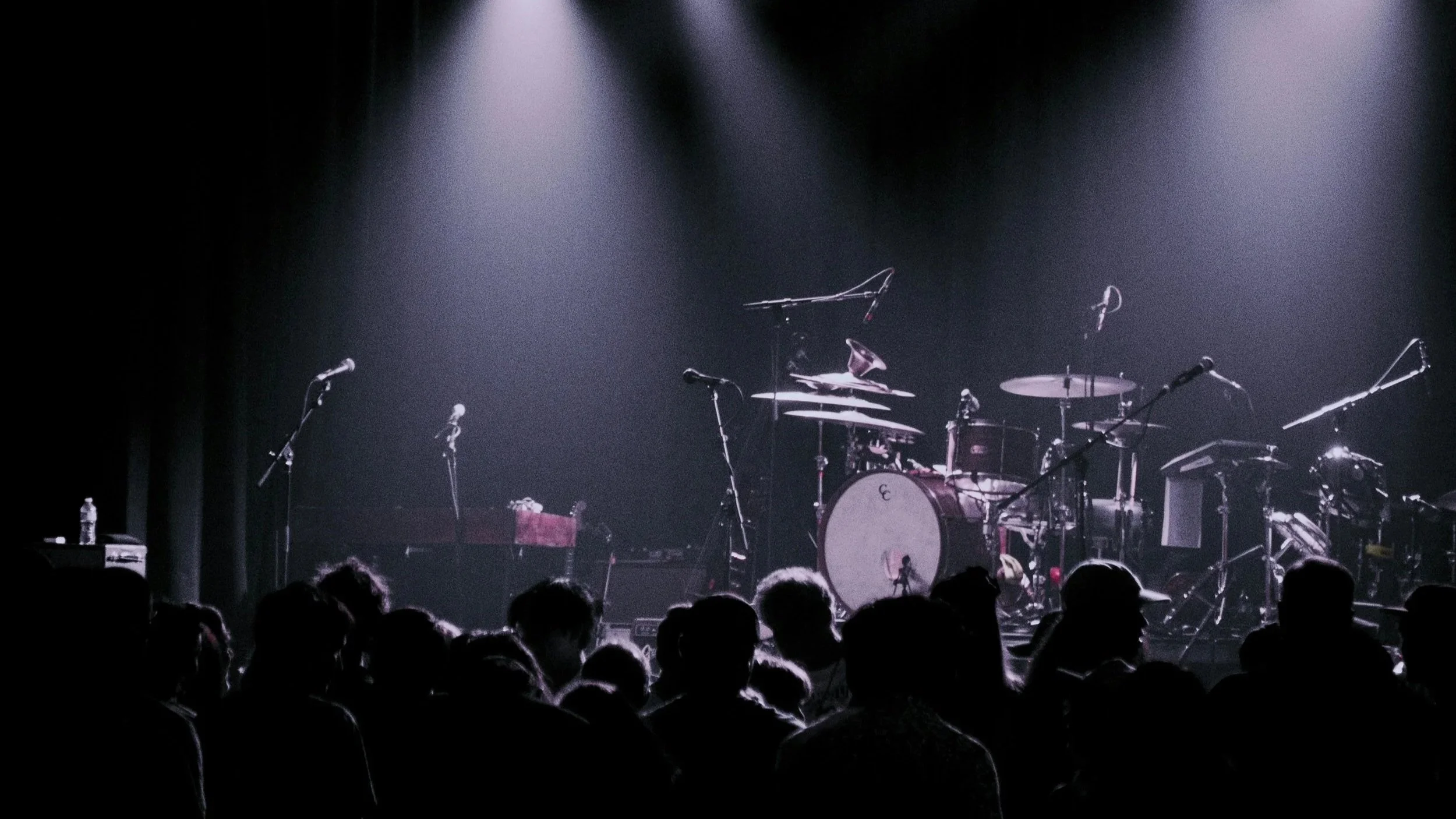 Scène de concert vide avec une scène éclairée par des projecteurs, comprenant une batterie et plusieurs micros, avec le public assis devant.