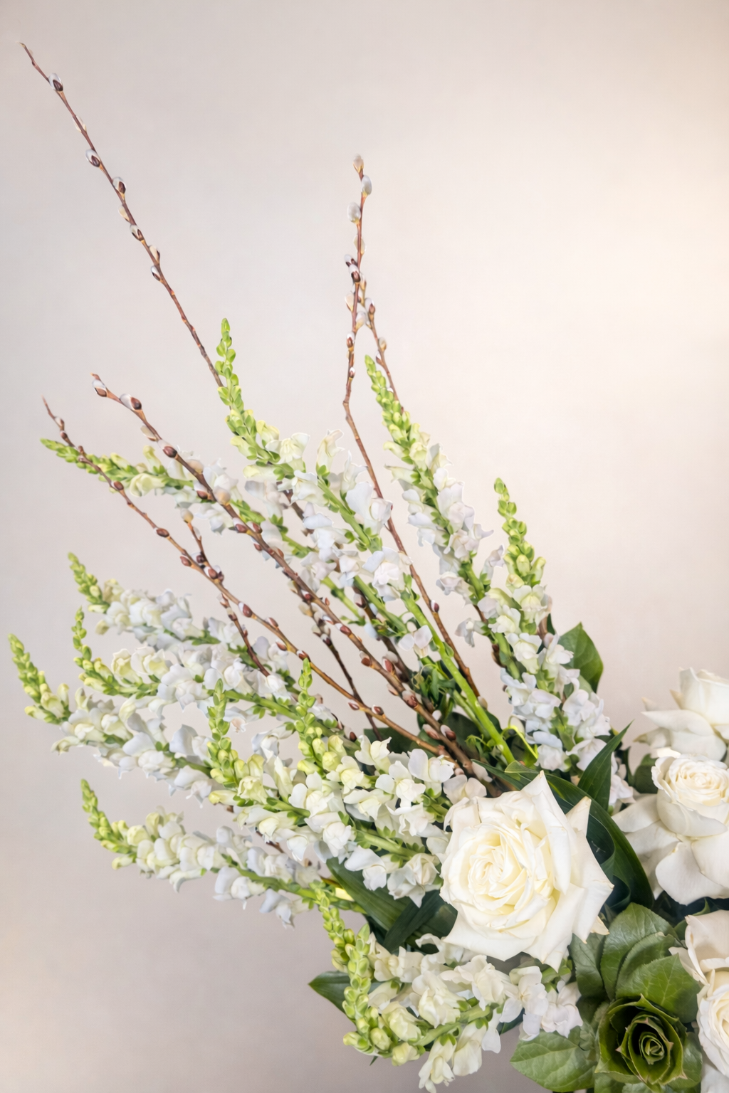 A floral arrangement of white roses, white snapdragons, and pussy willow branches against a plain background.