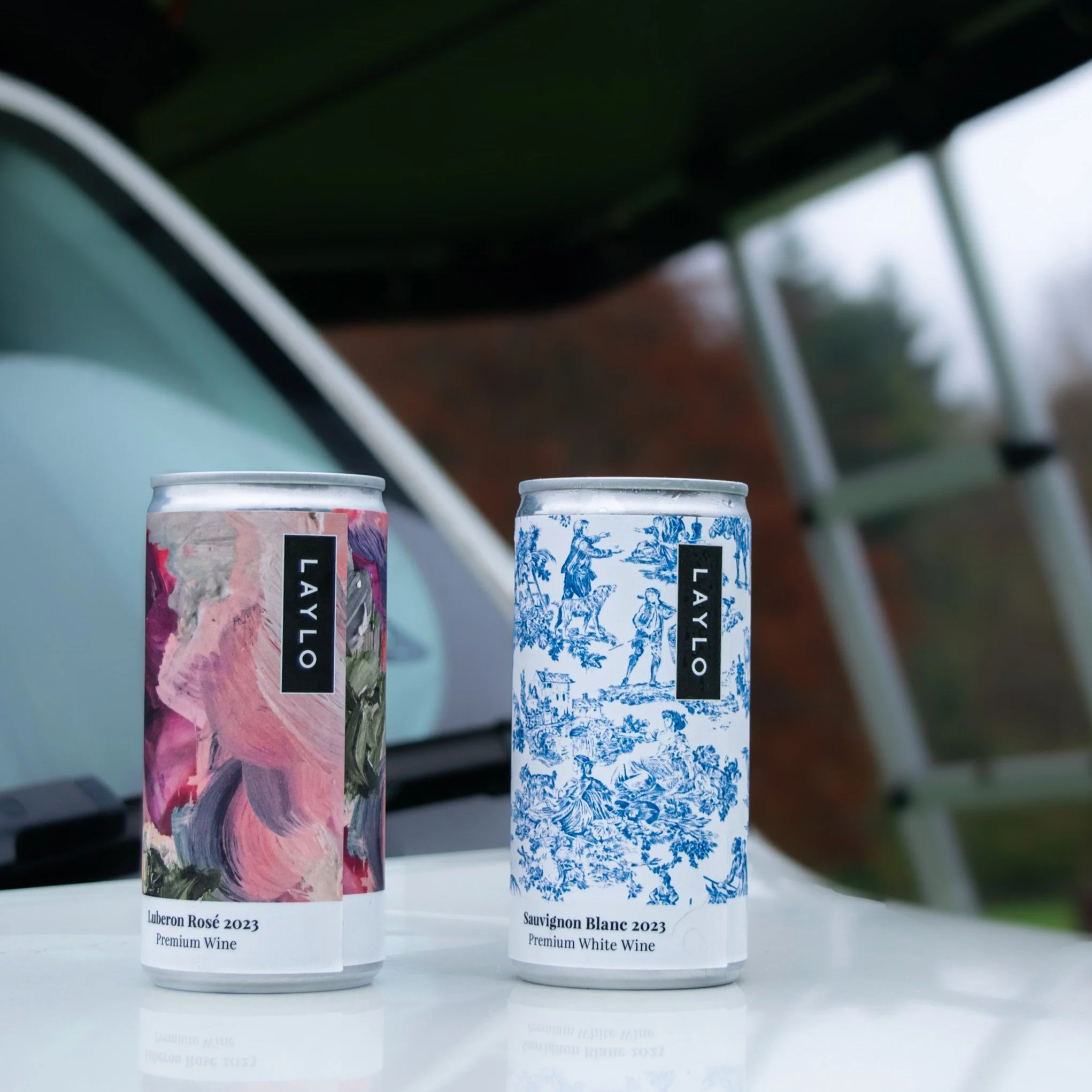 Two cans of wine, Luberon Rosé 2023 and Sauvignon Blanc 2023, placed on a reflective surface outdoors, with a blurred background of a roof top tent and greenery.