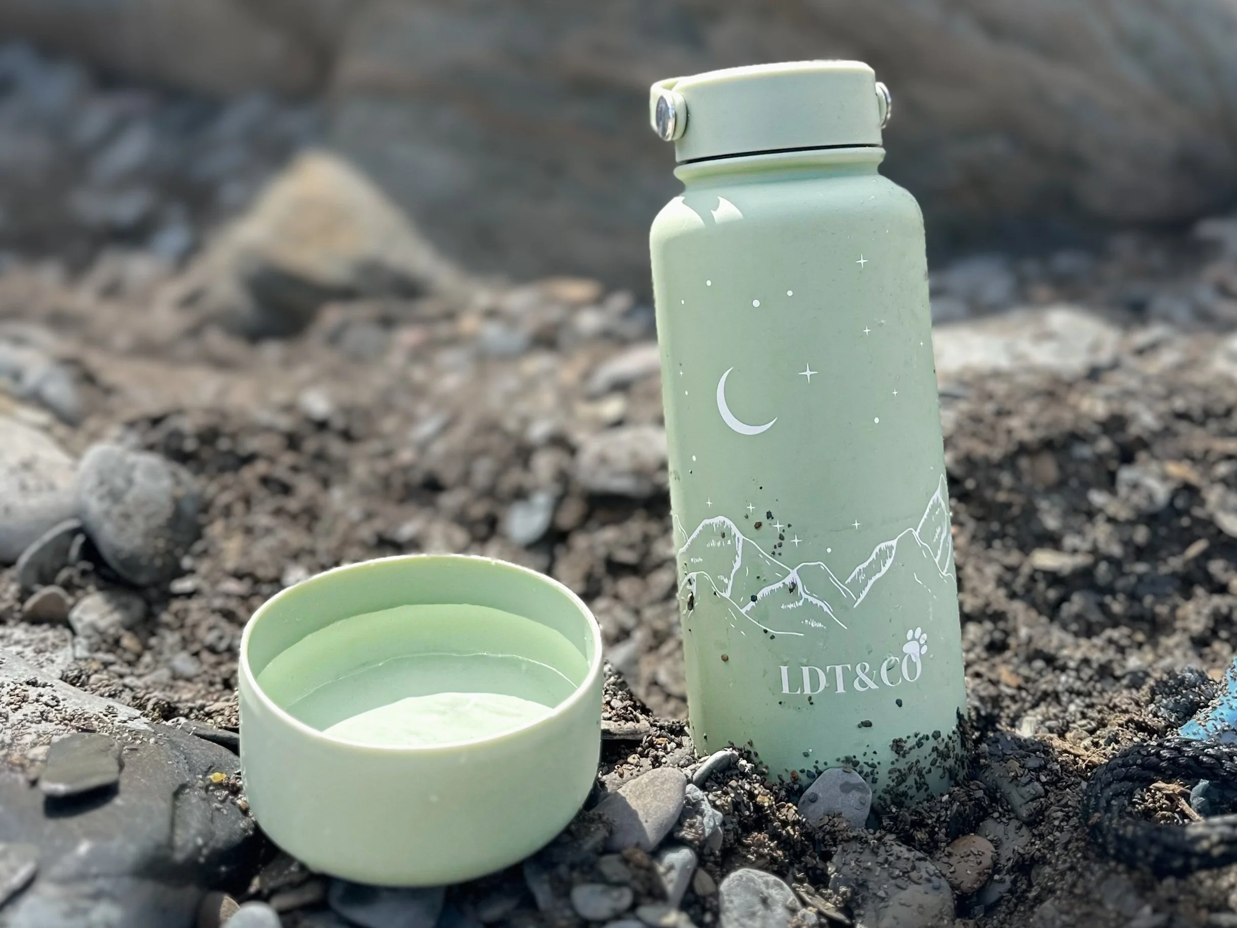 A light green water bottle with a celestial design featuring a moon and stars, standing upright on rocky ground. An open matching lid is placed in front of the bottle.